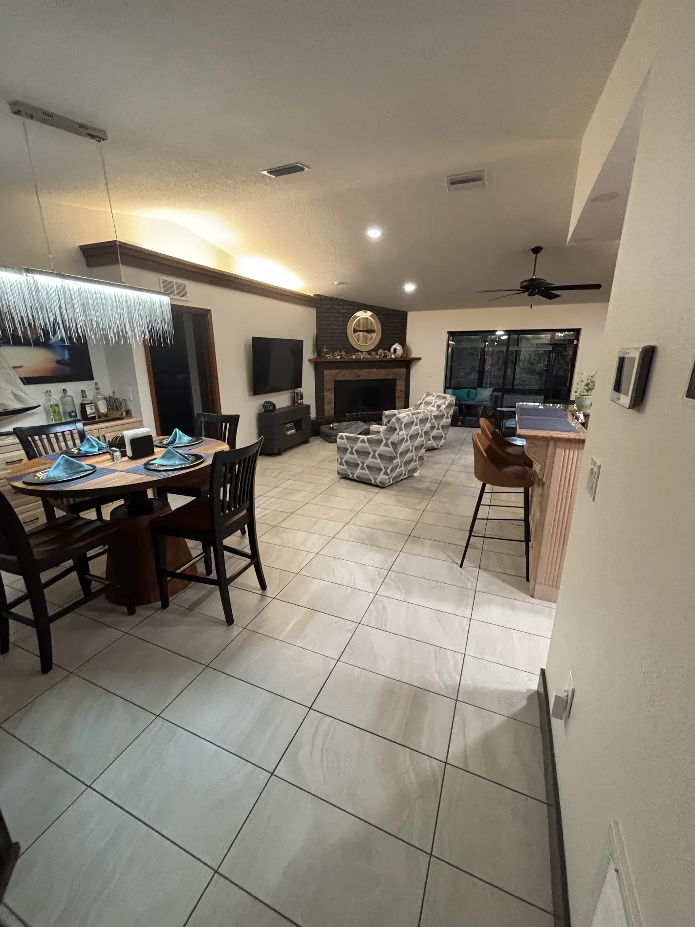 A living room and dining area with tiled floors, a fireplace with a brick surround, a wall-mounted TV, a dining table set with blue napkins, and various chairs, including bar stools, under warm ceiling lighting and a ceiling fan.