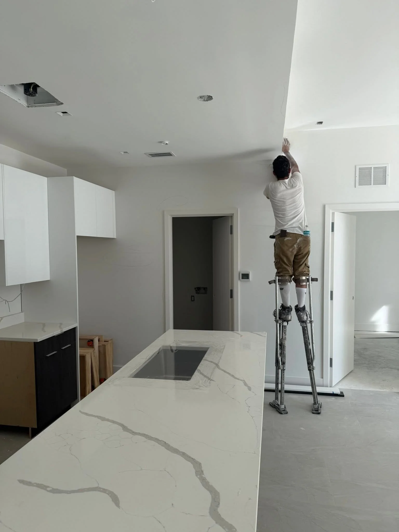 A person on a ladder installing or working on the ceiling of a modern kitchen, with white cabinets and a large marble countertop with gray veining.