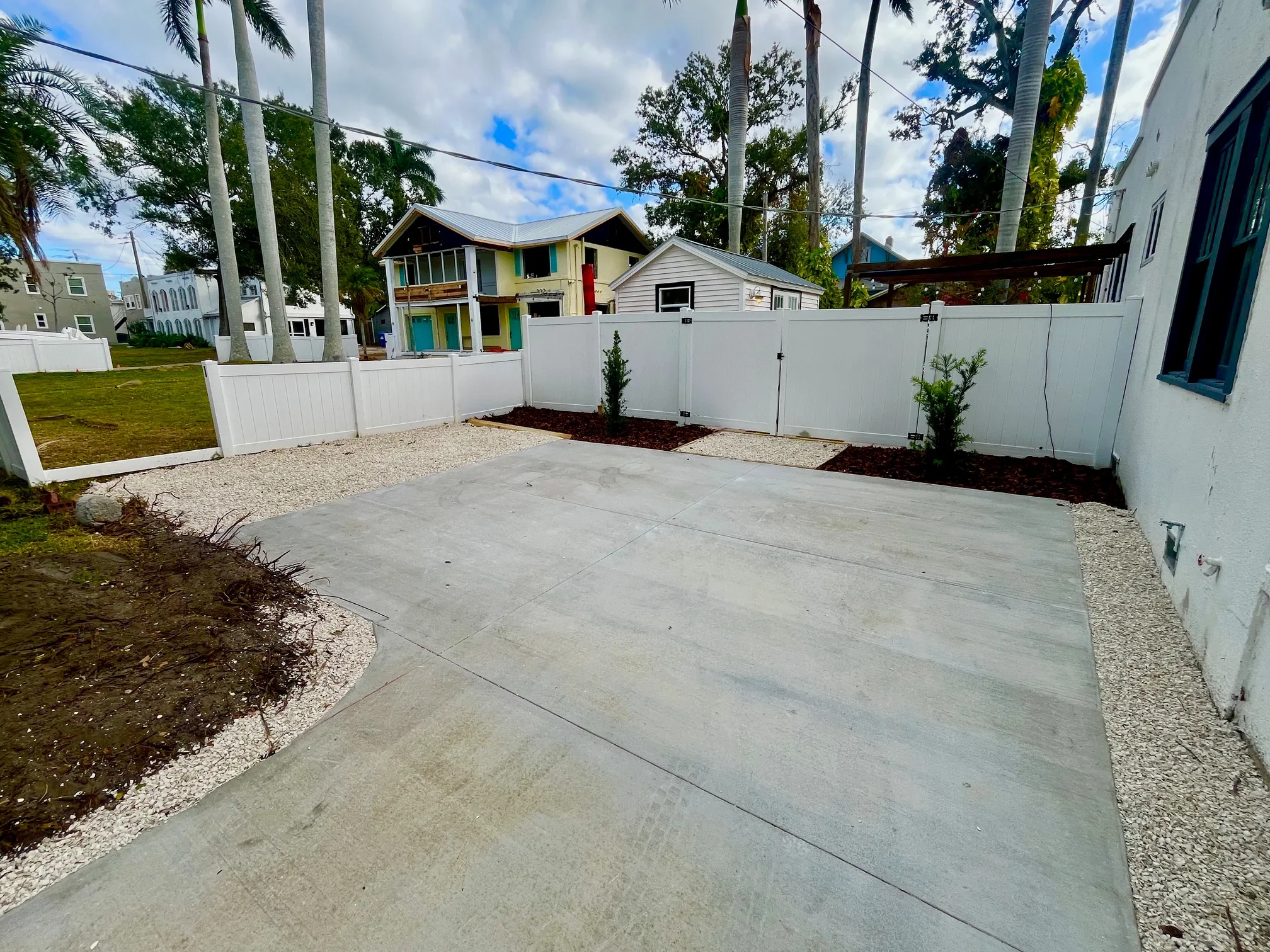 A backyard with a concrete patio, a white fence, small bushes, and houses in the background under a partly cloudy sky, with tall palm trees and other trees visible.