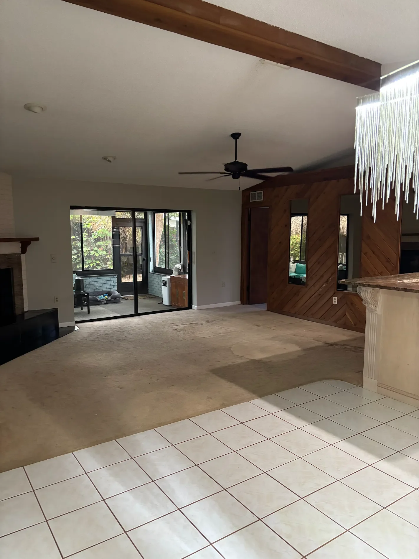Interior of a living space with a beige carpeted floor, white walls, wooden wall paneling, a ceiling fan, and a sliding glass door leading to an outdoor area with trees and plants.