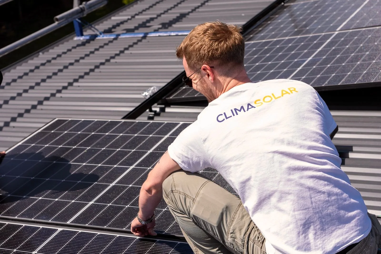 A man working on solar panels on a rooftop, wearing a white shirt that says CLIMASOLAR in yellow and purple letters.