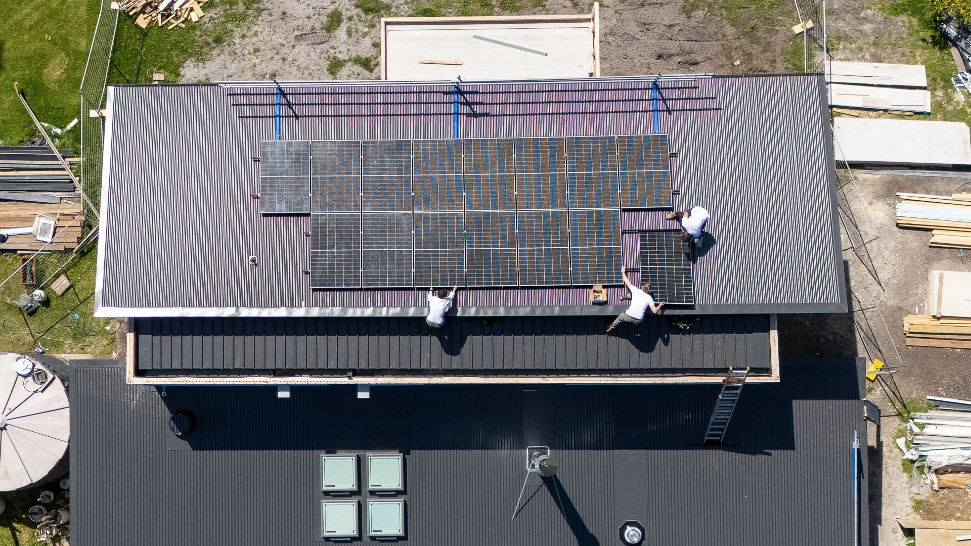 Three workers installing solar panels on a rooftop with other buildings and construction materials surrounding the area.