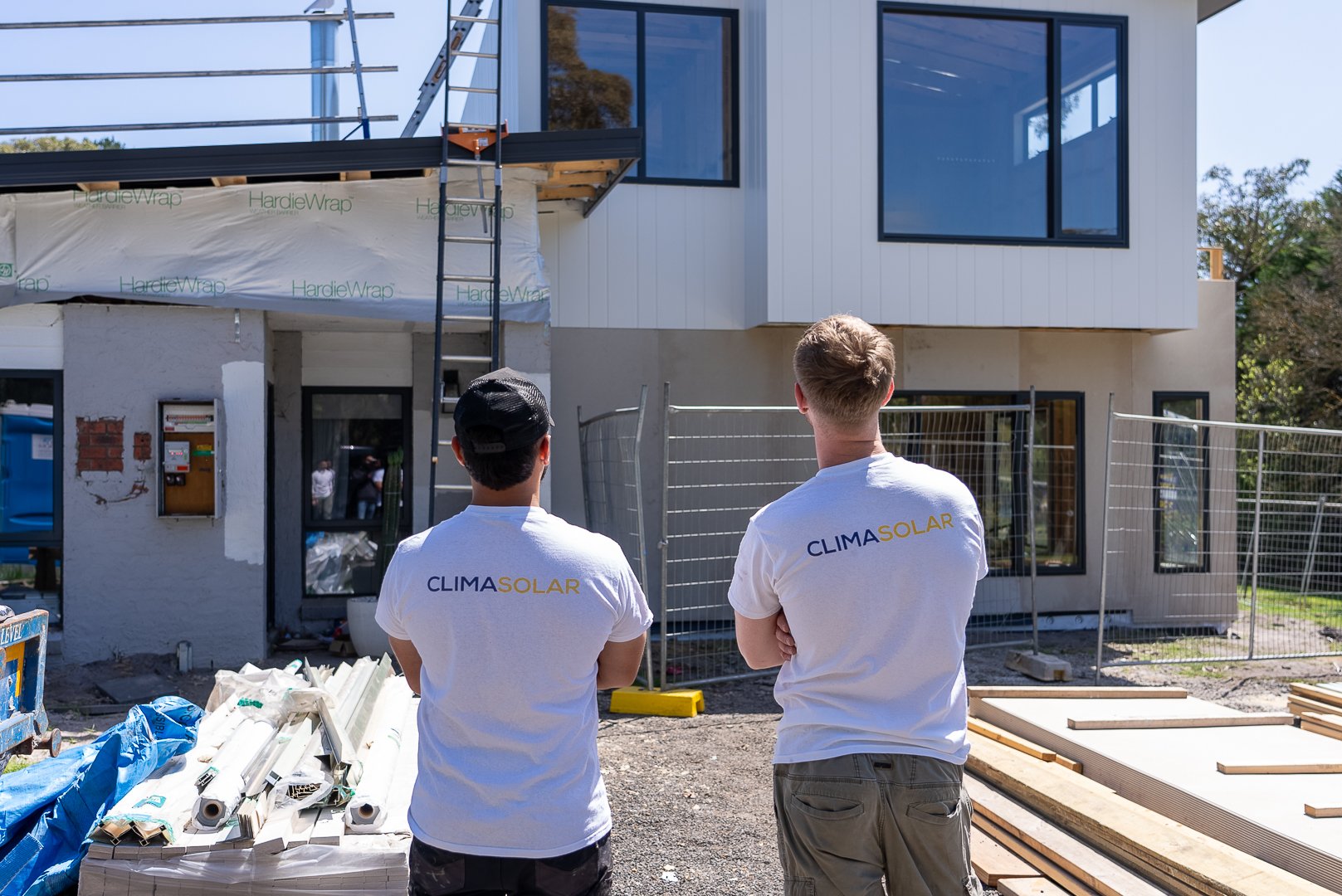 Two workers with CLIMASOLAR shirts standing in front of a modern house under construction, observing the building site.