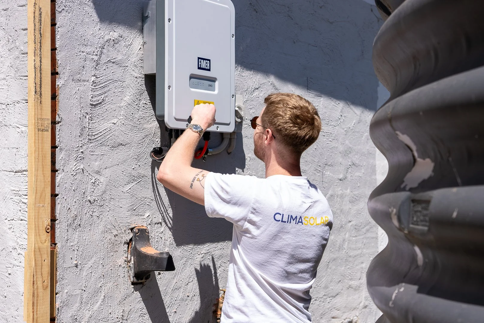A man with glasses and a white T-shirt with 'CLIMASOLAR' printed on the back is working on an electrical panel attached to a wall outside. He's adjusting or inspecting the panel on a sunny day.