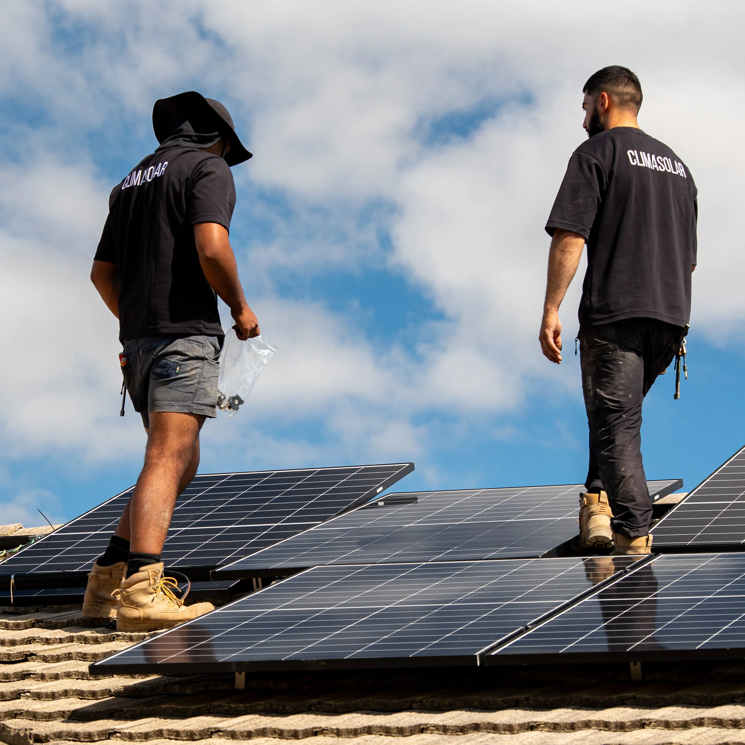 Two workers installing solar panels on a roof under a partly cloudy sky.