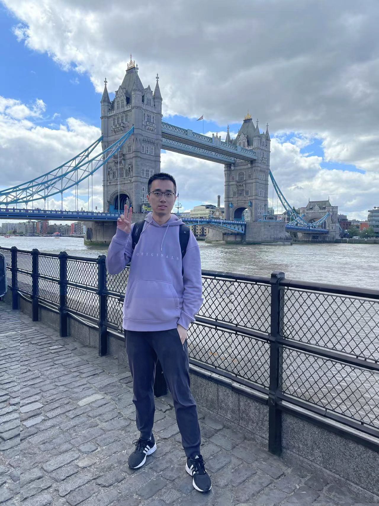 Young man standing on a cobblestone path near the Thames River in London, making a peace sign with his right hand, with Tower Bridge in the background under a partly cloudy sky.