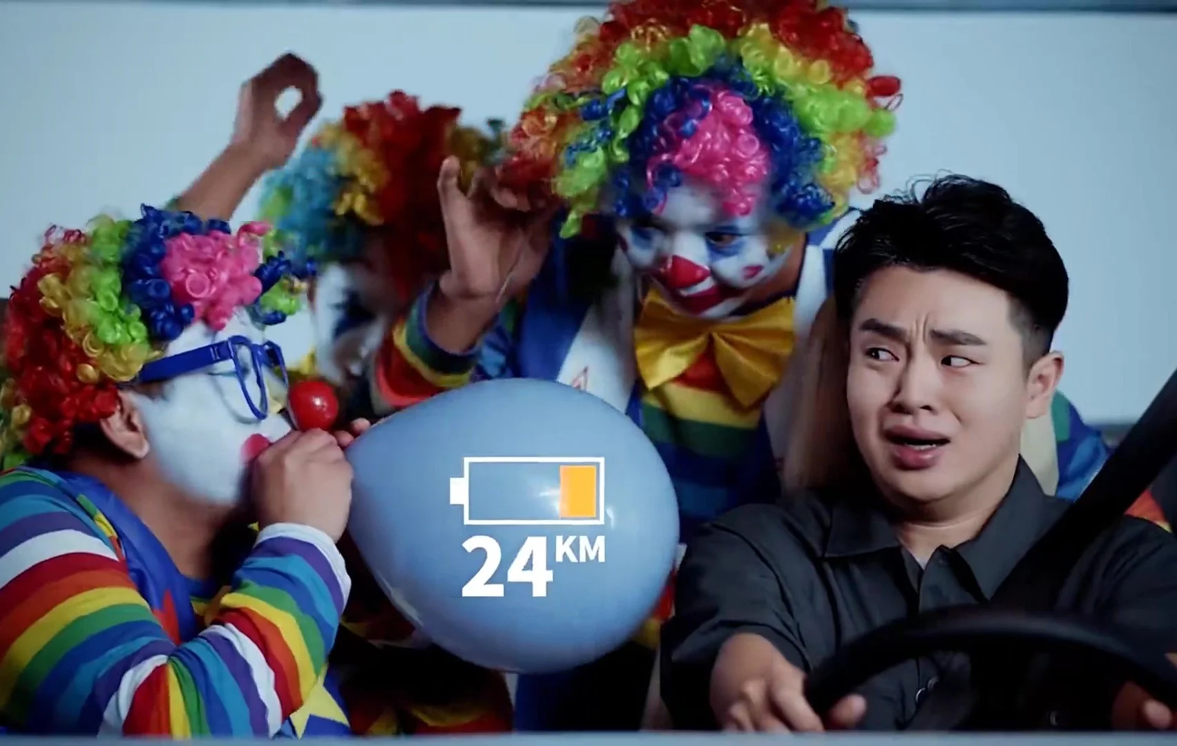 A man sitting in a car looks worried as three clowns with colorful wigs, white face paint, and clown costumes surround him. One clown is blowing up a balloon labeled '24 KM'.