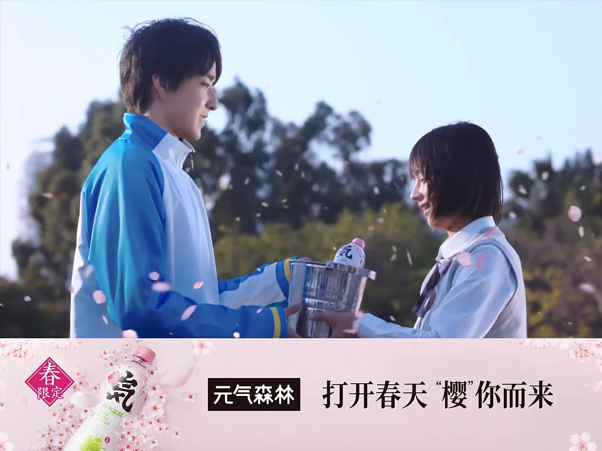 A young male and female exchange a drink outdoors during spring, with cherry blossom petals falling around them. The scene suggests a friendly or romantic moment.