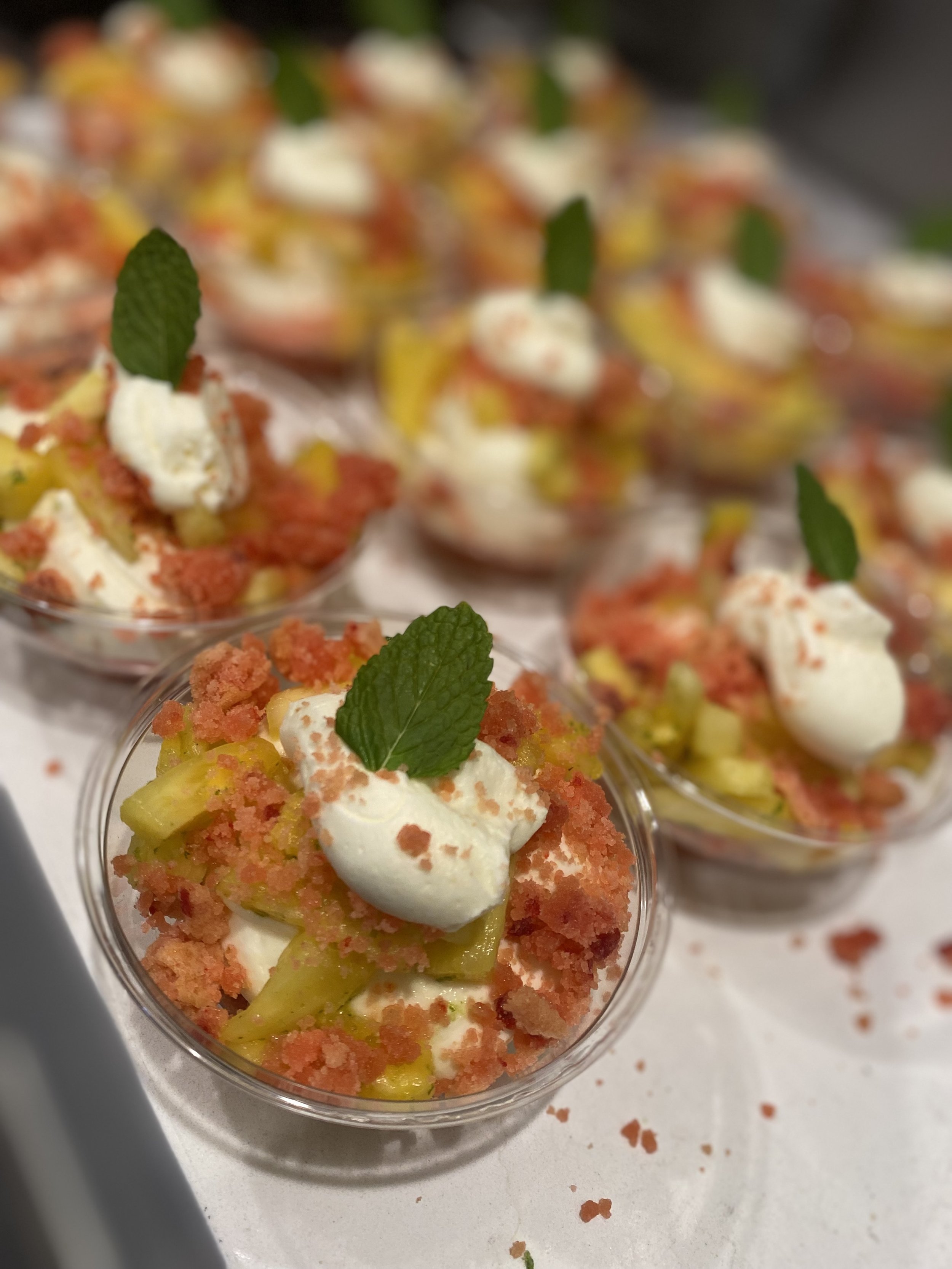 Individual serving cups of fruit crumble topped with whipped cream and a mint leaf, garnished with red crumbs.