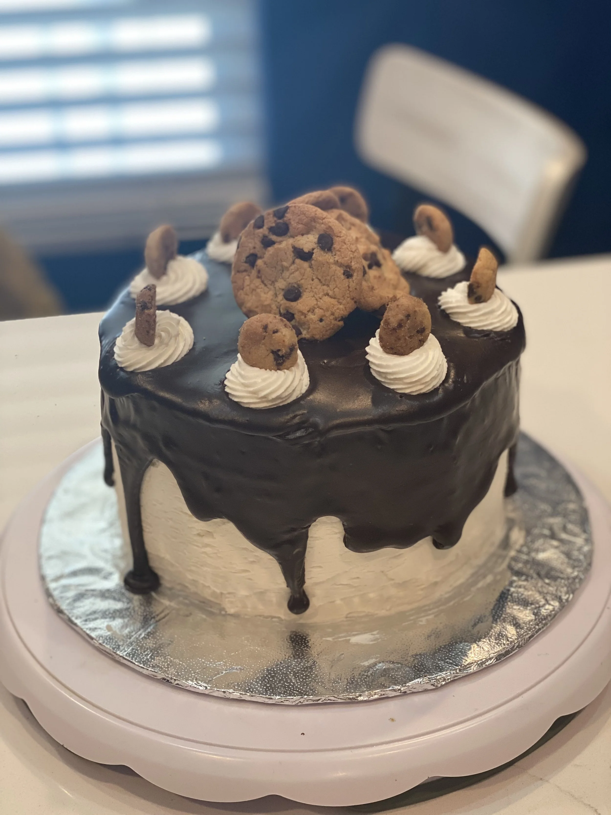 Chocolate cake with chocolate ganache, decorated with white frosting, mini cookies, and chocolate chip cookies on top, placed on a silver cake board.