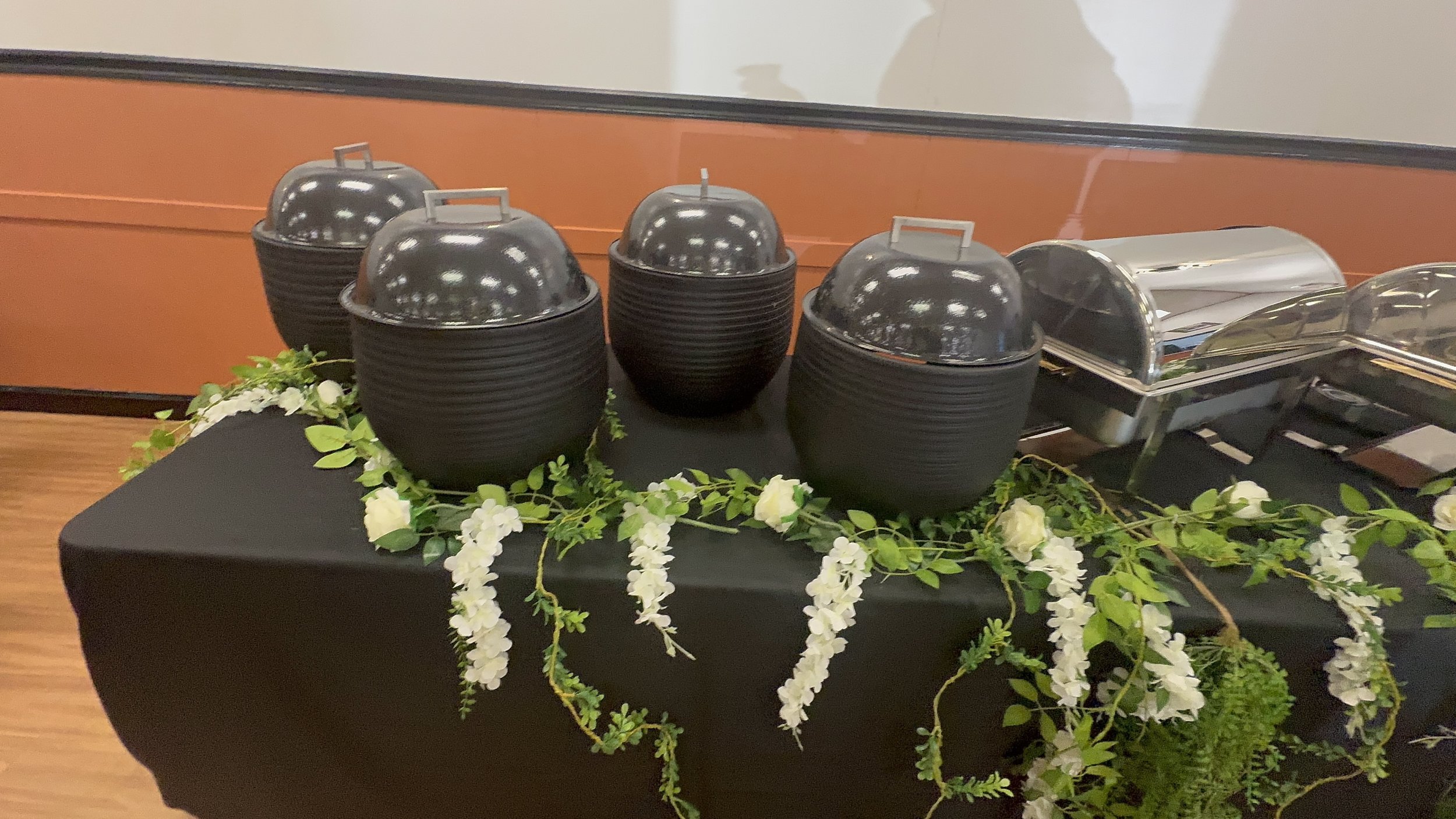 Black bowls with silver lids on a table decorated with white flowers and green foliage, with metal chafing dishes in the background.