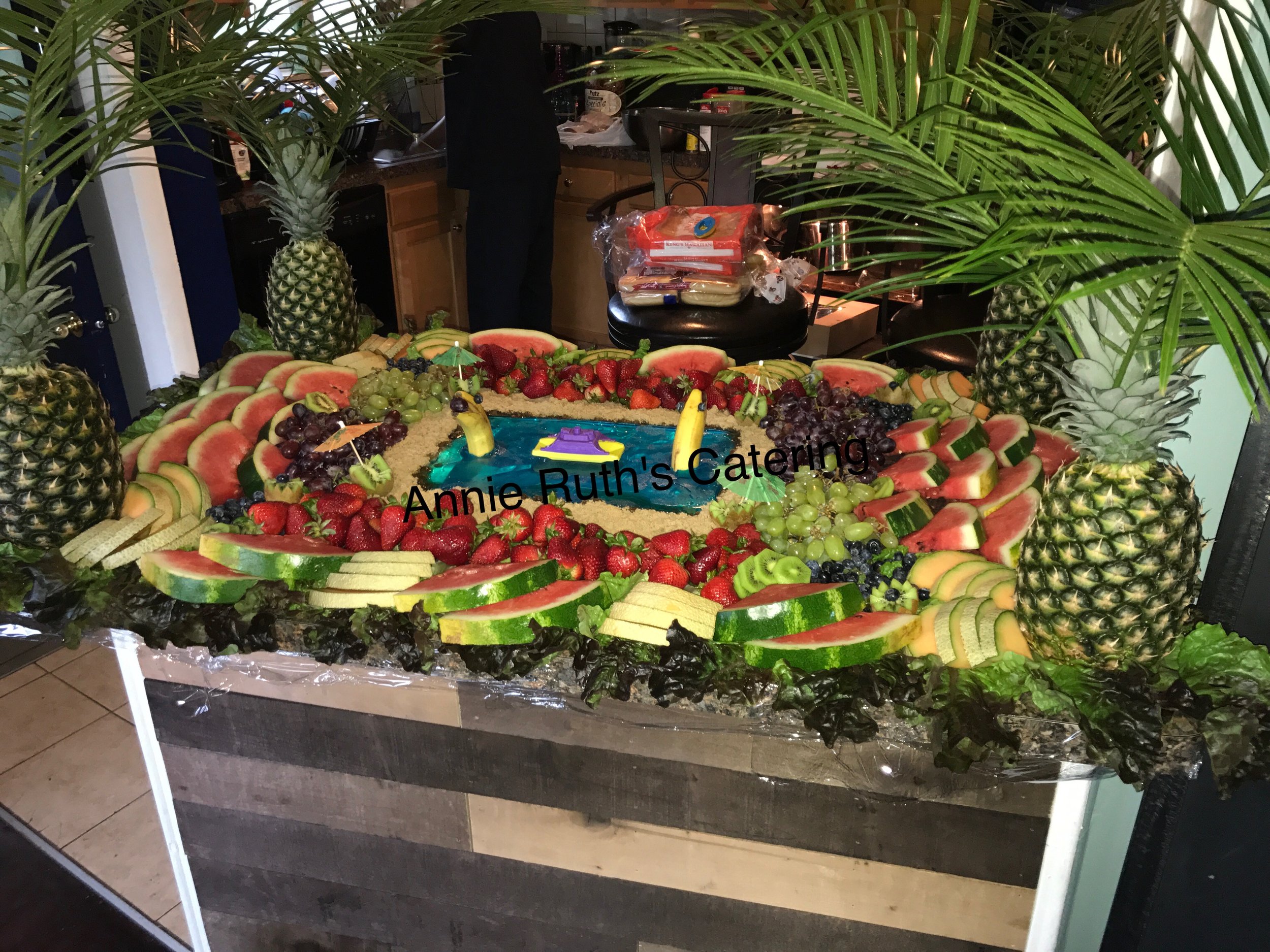 A large display of fresh sliced fruits arranged around a small pond with toy boat, including pineapples, watermelons, cantaloupe, strawberries, grapes, and kiwi, with a sign reading "Annie Ruth's Catering".