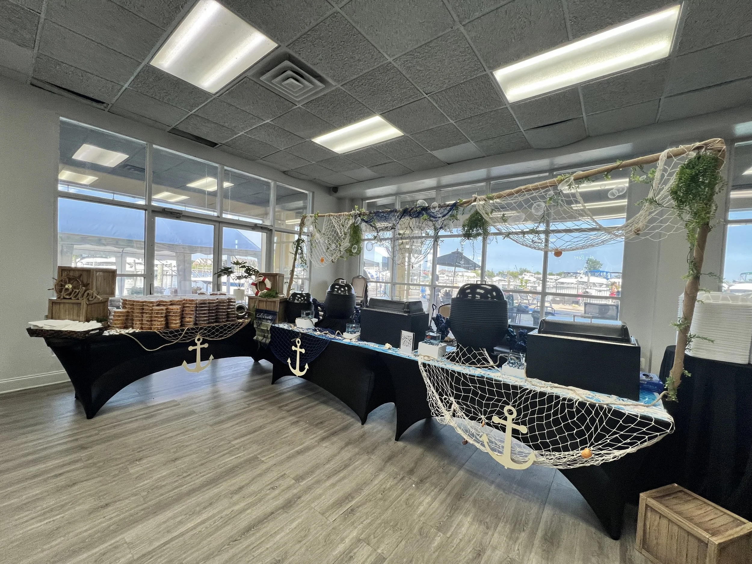 A sea-themed event setup with a table decorated with nautical ornaments, including anchor cutouts, netting, and greenery, with food items stacked on one side, inside a glass room with large windows and marina view.