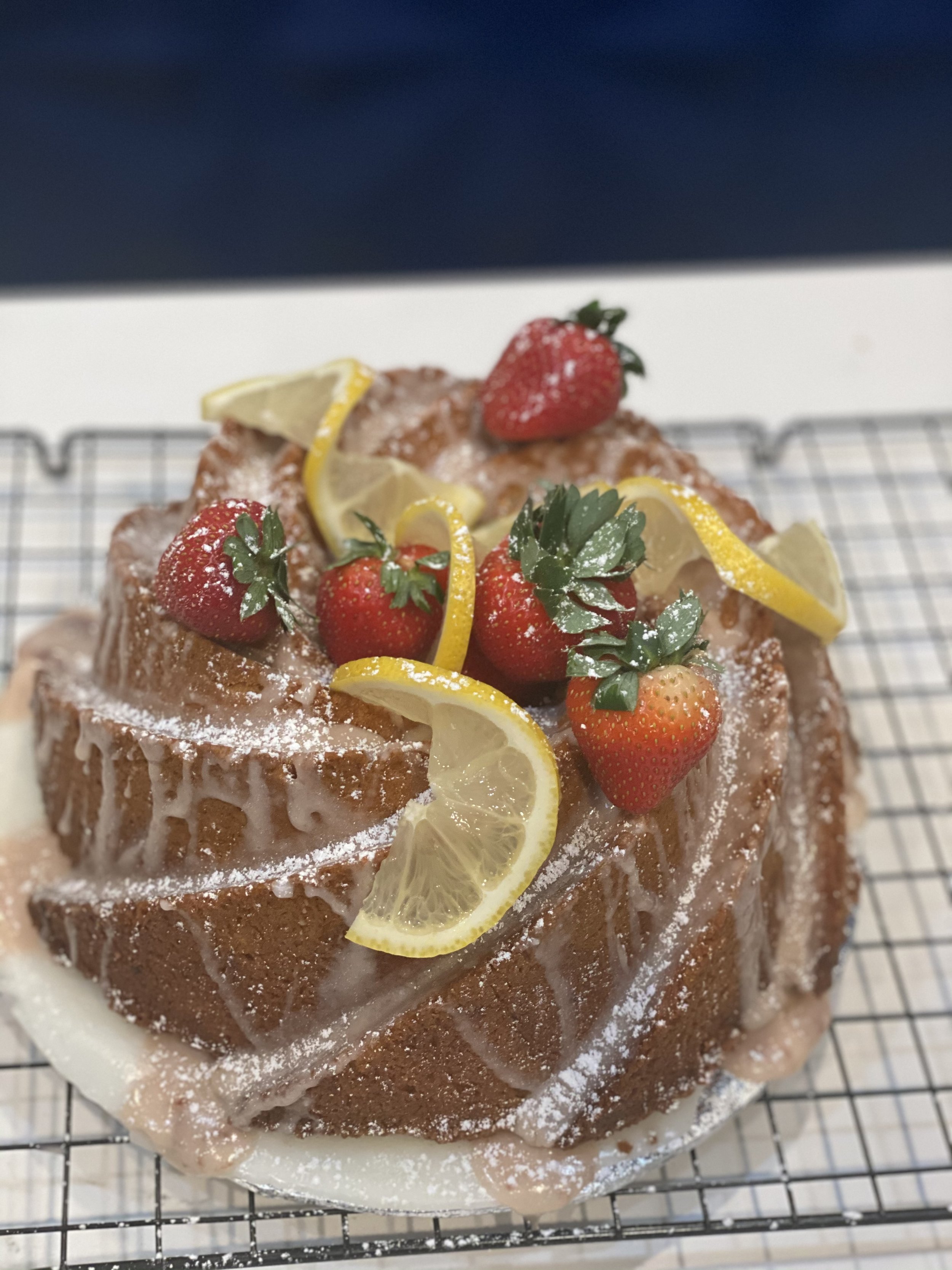 A lemon drizzle cake topped with strawberries, lemon slices, and powdered sugar, displayed on a wire rack.