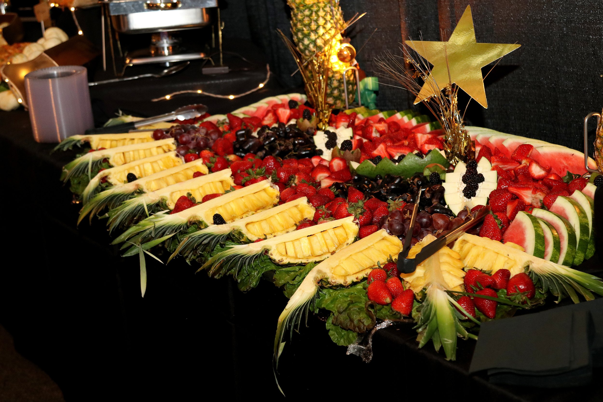 A large fruit platter featuring pineapple slices, watermelon wedges, strawberries, grapes, blackberries, and other assorted fruits, decorated with gold star-shaped ornaments and gold tinsel.