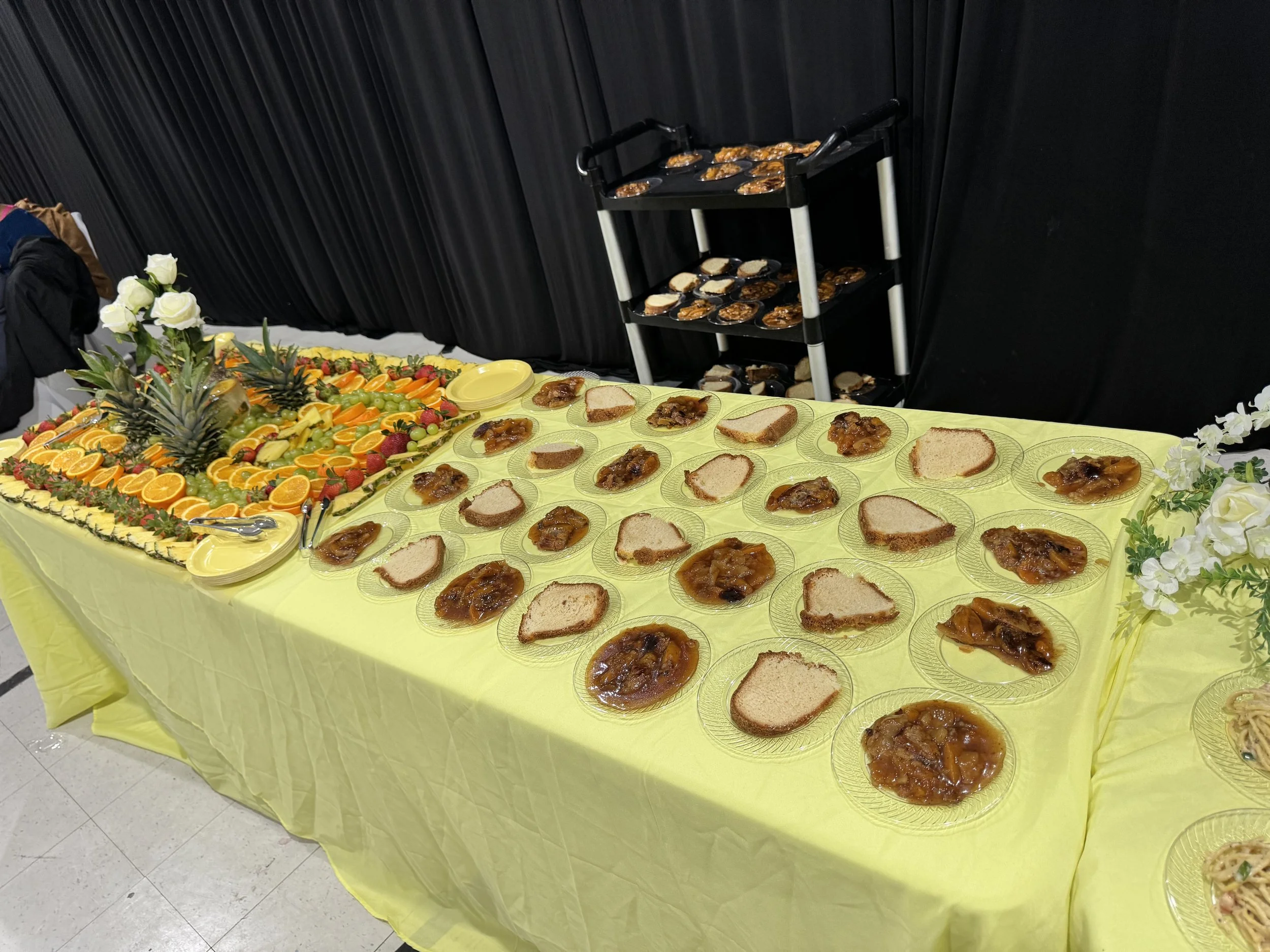 Buffet table with sliced bread, fruit, and fruit preserves or jam in bowls, decorated with flowers, in an event setting.