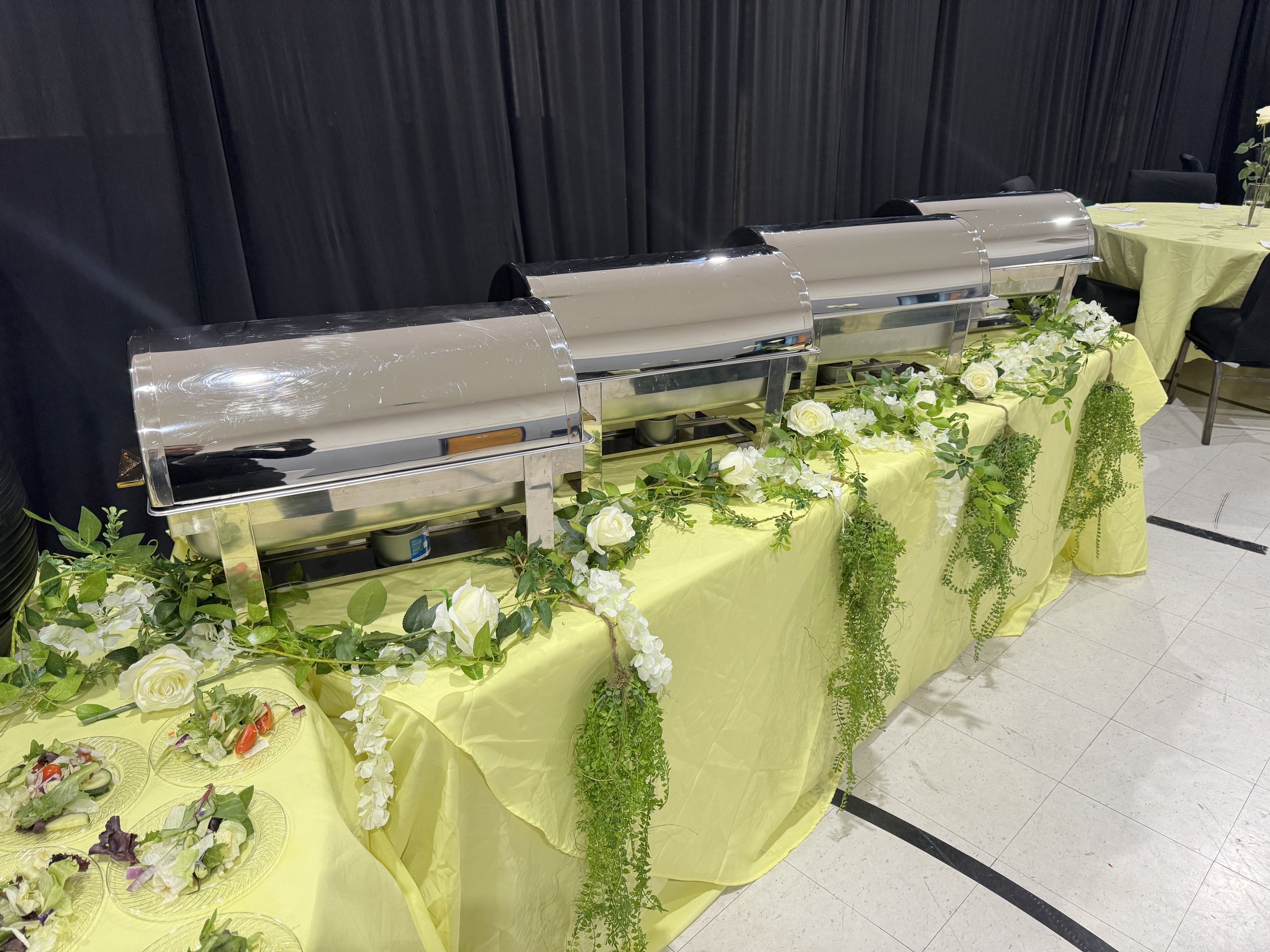 Buffet table with chafing dishes, decorated with white flowers and green foliage, covered with a yellow tablecloth in a banquet setting.