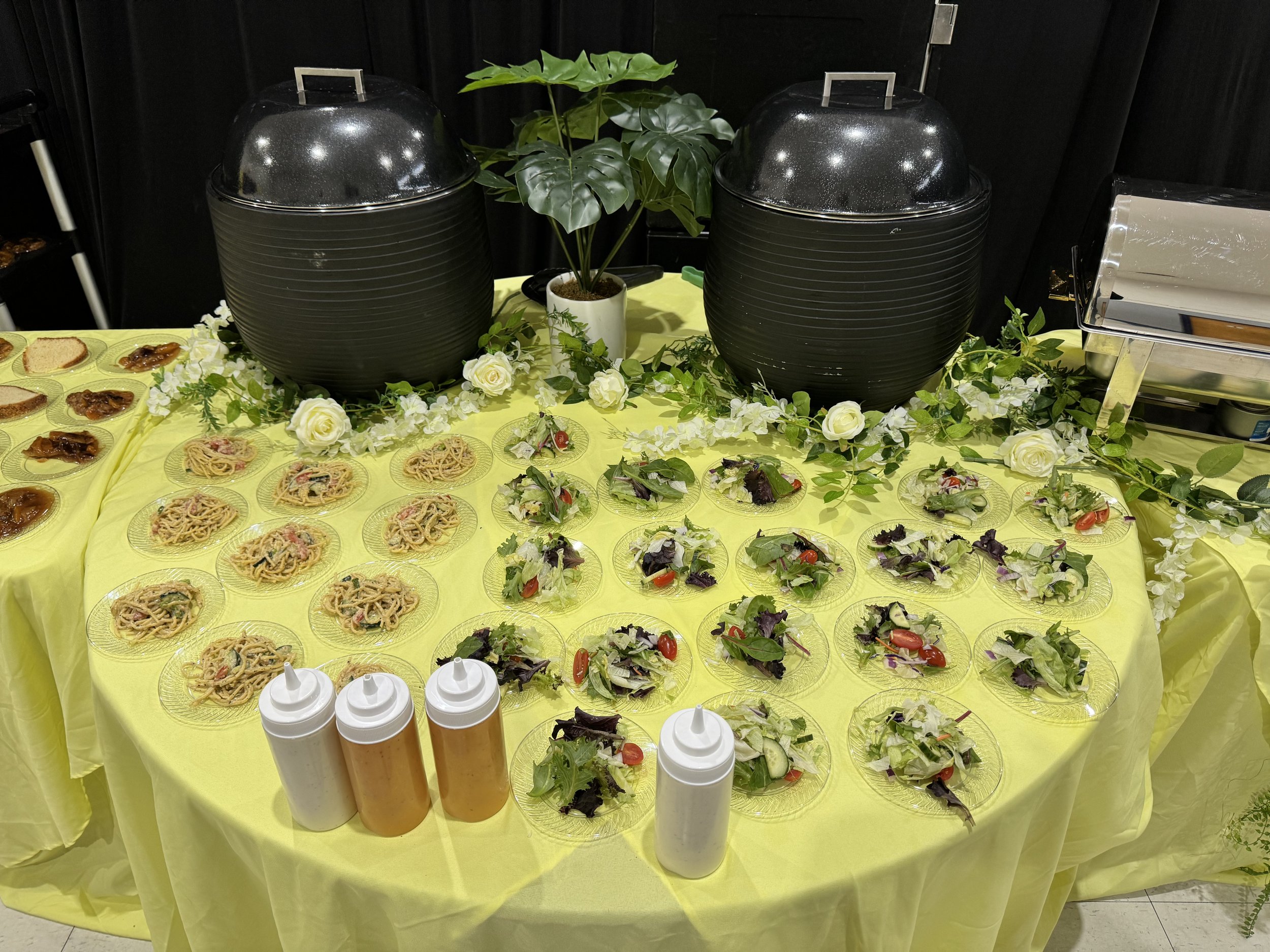 A buffet table with salad, pasta, and sauces, decorated with white roses and green leaves, with two large black containers and a potted plant in the background.