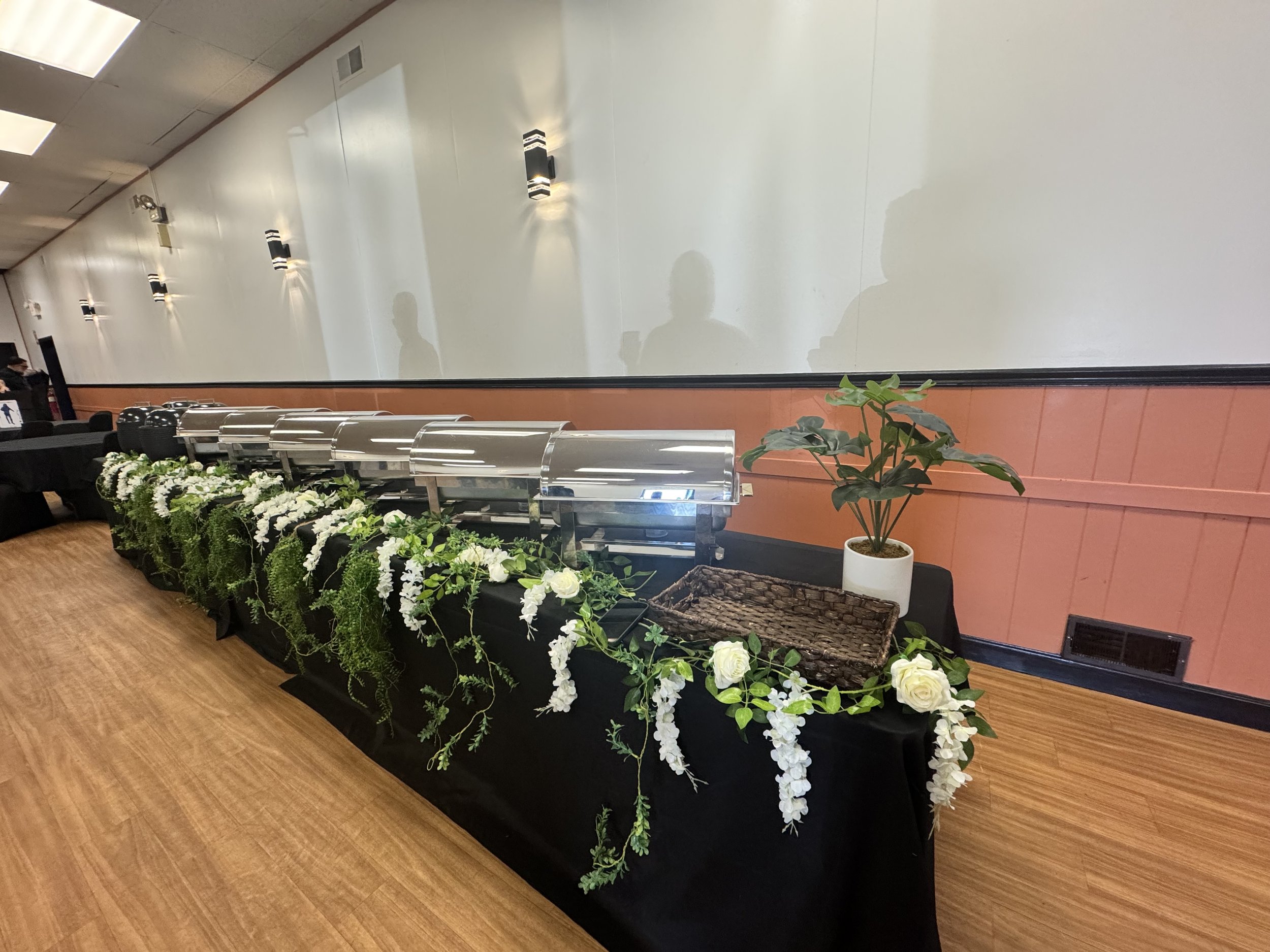 A buffet table decorated with white flowers and greenery, featuring chafing dishes and a potted plant, set against a wall with black trim and wood flooring.