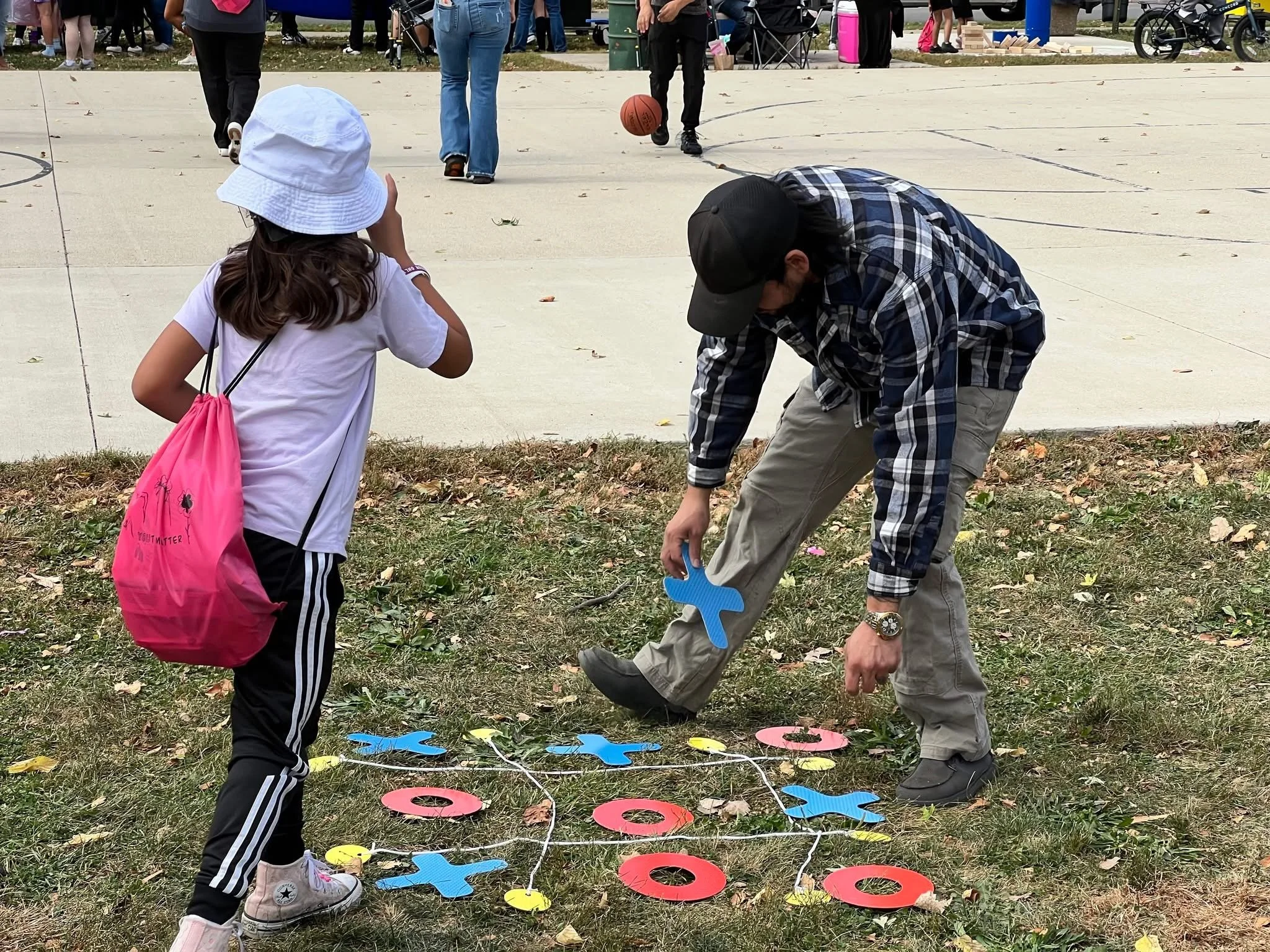 Children and adults playing outdoor Twister game on grass and concrete with a crowd gathered in the background.