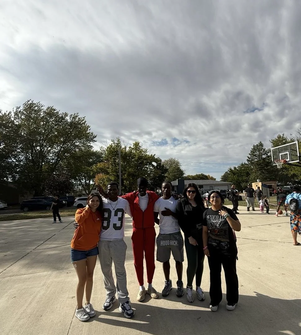 Group of six diverse teenagers standing together on an outdoor basketball court, with some holding each other, smiling and looking at the camera. In the background, other people are socializing, and a basketball hoop is visible.