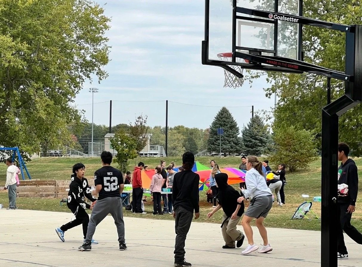 A group of children and teenagers playing basketball on an outdoor court, with some people in the background preparing for a picnic or event under colorful umbrellas.