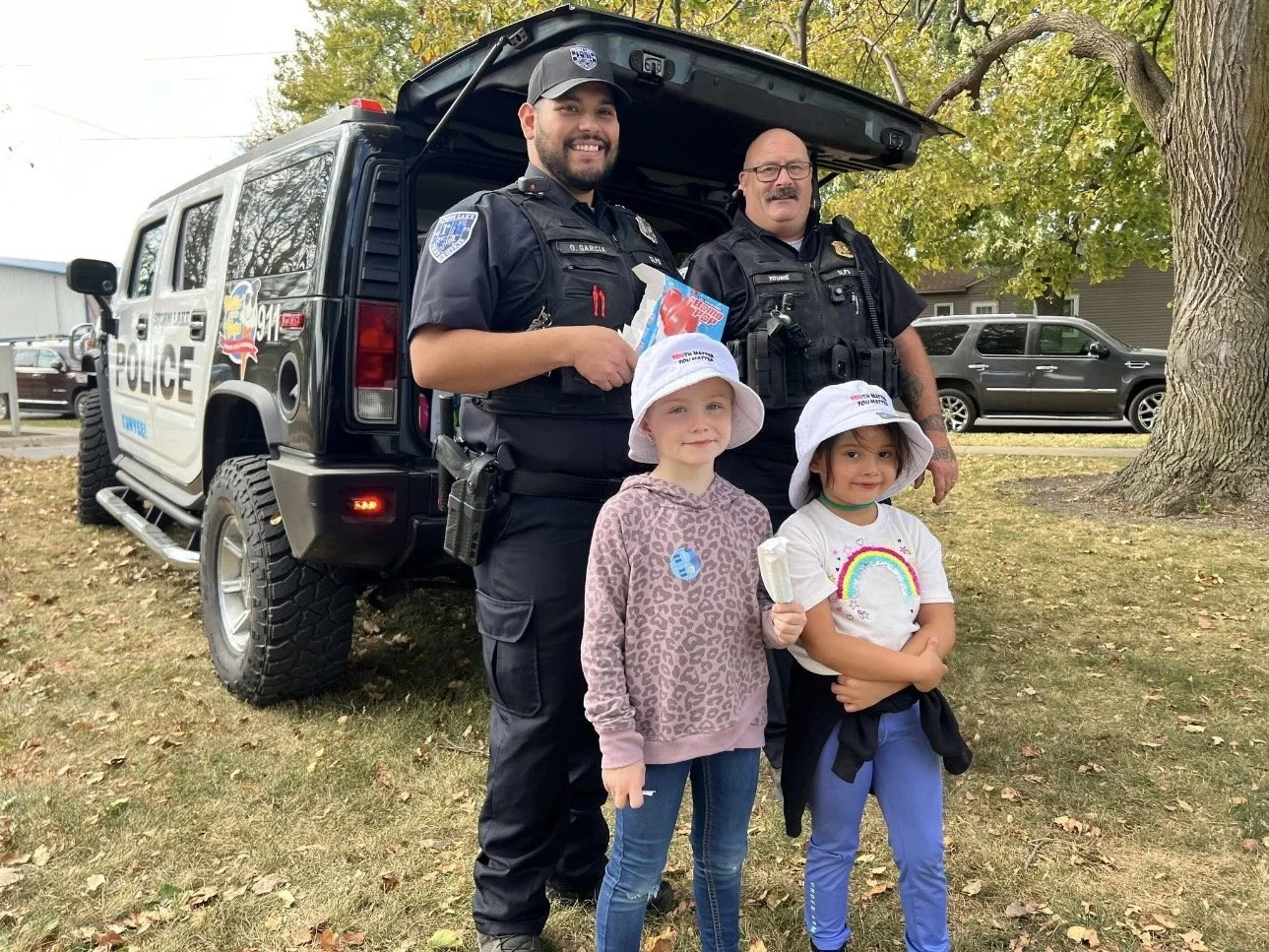 Two police officers standing behind two young girls holding ice pops during an outdoor community event, with police vehicle, cars, trees, and a residential building in the background.