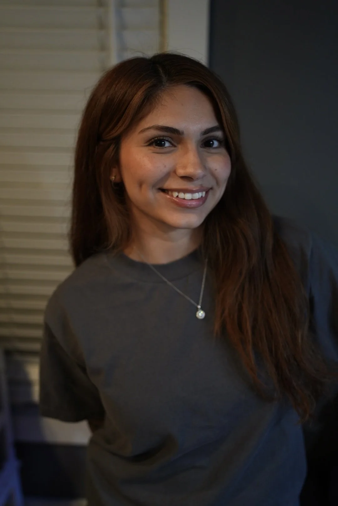 A young woman with long brown hair smiling, wearing a dark gray shirt and a silver necklace with a pendant, standing indoors.