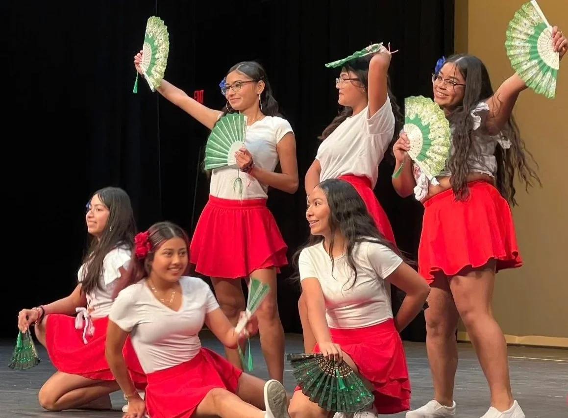 Group of six young women performing a dance on stage, dressed in white tops and red skirts, holding green fans, some standing and others kneeling.