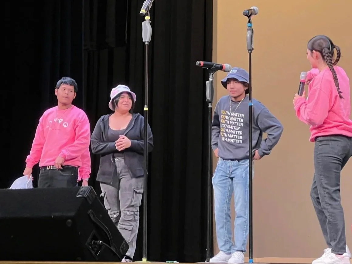 Four young people on stage participating in a discussion or performance, with three microphones, two girls and two boys, one girl holds a microphone, and a black curtain in the background.