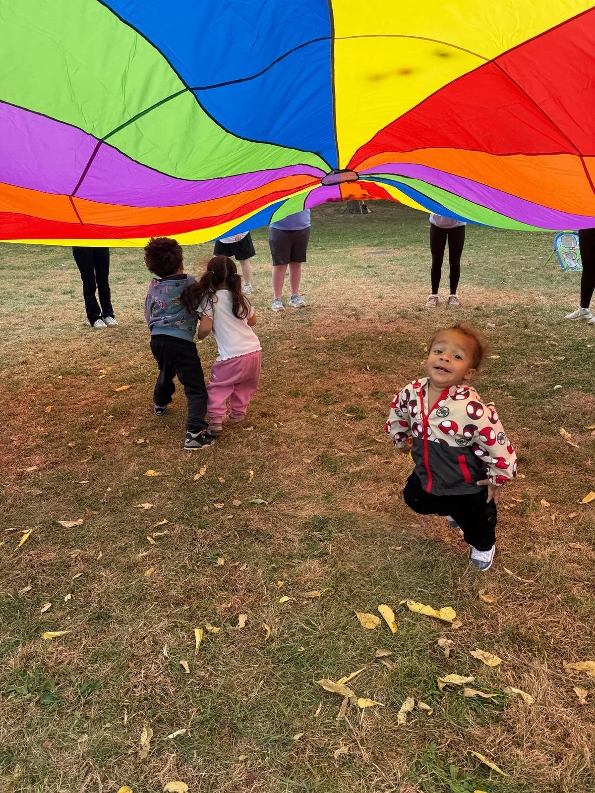 Children playing with a colorful parachute outdoors, with one young boy in the foreground smiling. The parachute is spread out overhead, and the children are holding onto the edges while standing on a grassy area with scattered leaves.