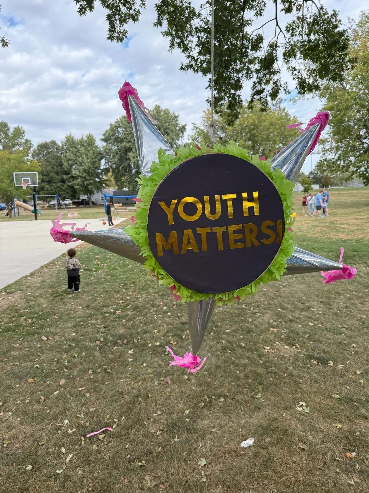 A festive balloon with a sign that says 'YOUTH MATTERS!' in gold letters, decorated with pink and green paper and hanging in a park with children and adults engaging in outdoor activities.