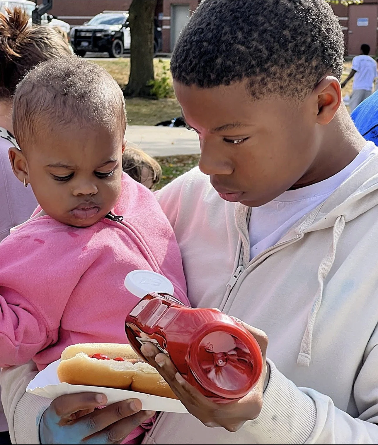 A young man holding a girl and a plate with a hot dog and a bottle of ketchup.