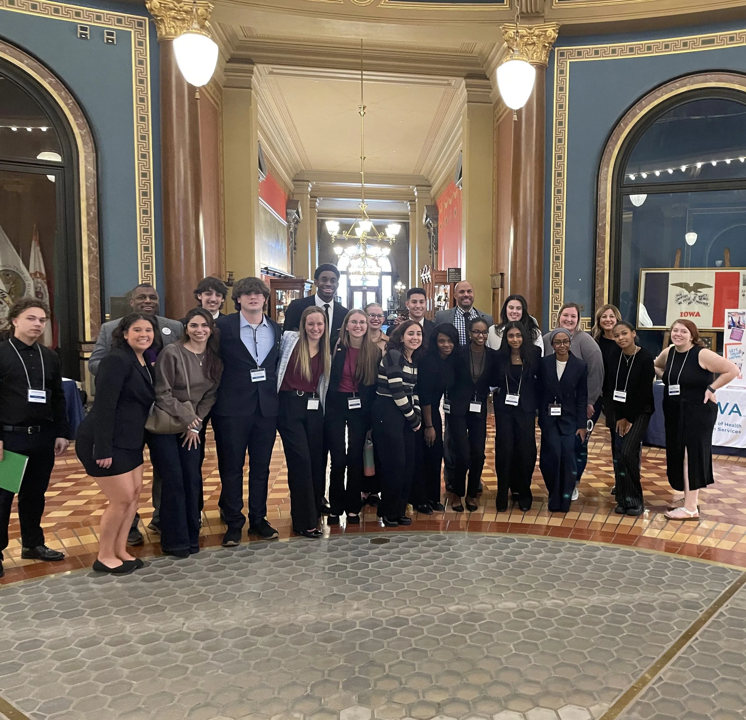 Group of people standing in a beautifully decorated hallway with ornate gold trimmings, chandeliers, and arched windows, smiling at the camera.