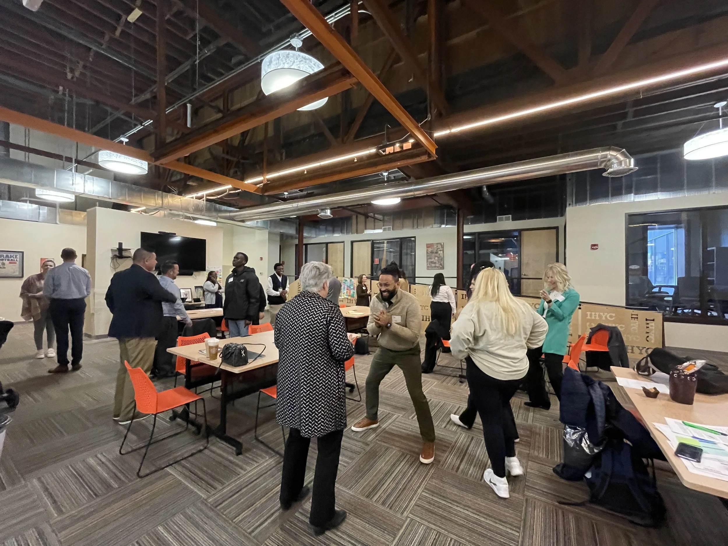 People gathered in a modern indoor space with high ceilings, wooden beams, and industrial-style ductwork, socializing at tables with orange chairs.
