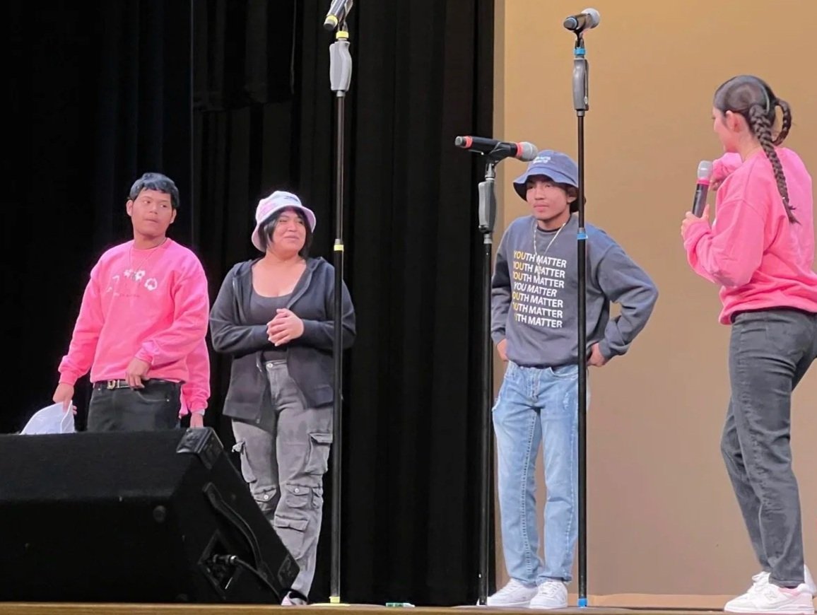 Four children stand on a stage with microphones, participating in a discussion or performance, with a black curtain background and a beige wall. One girl is speaking into a pink microphone.