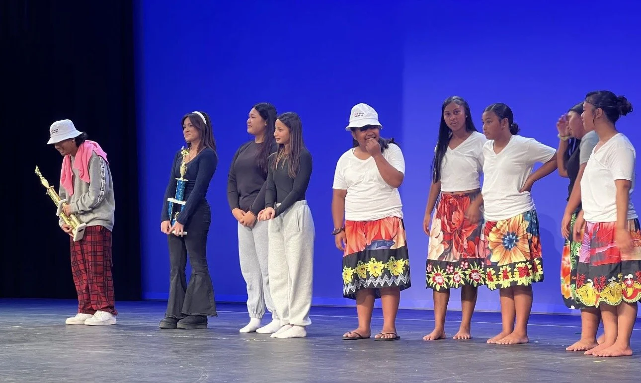 Group of girls standing on stage, some holding trophies, wearing casual and colorful dresses, with a plain blue background.