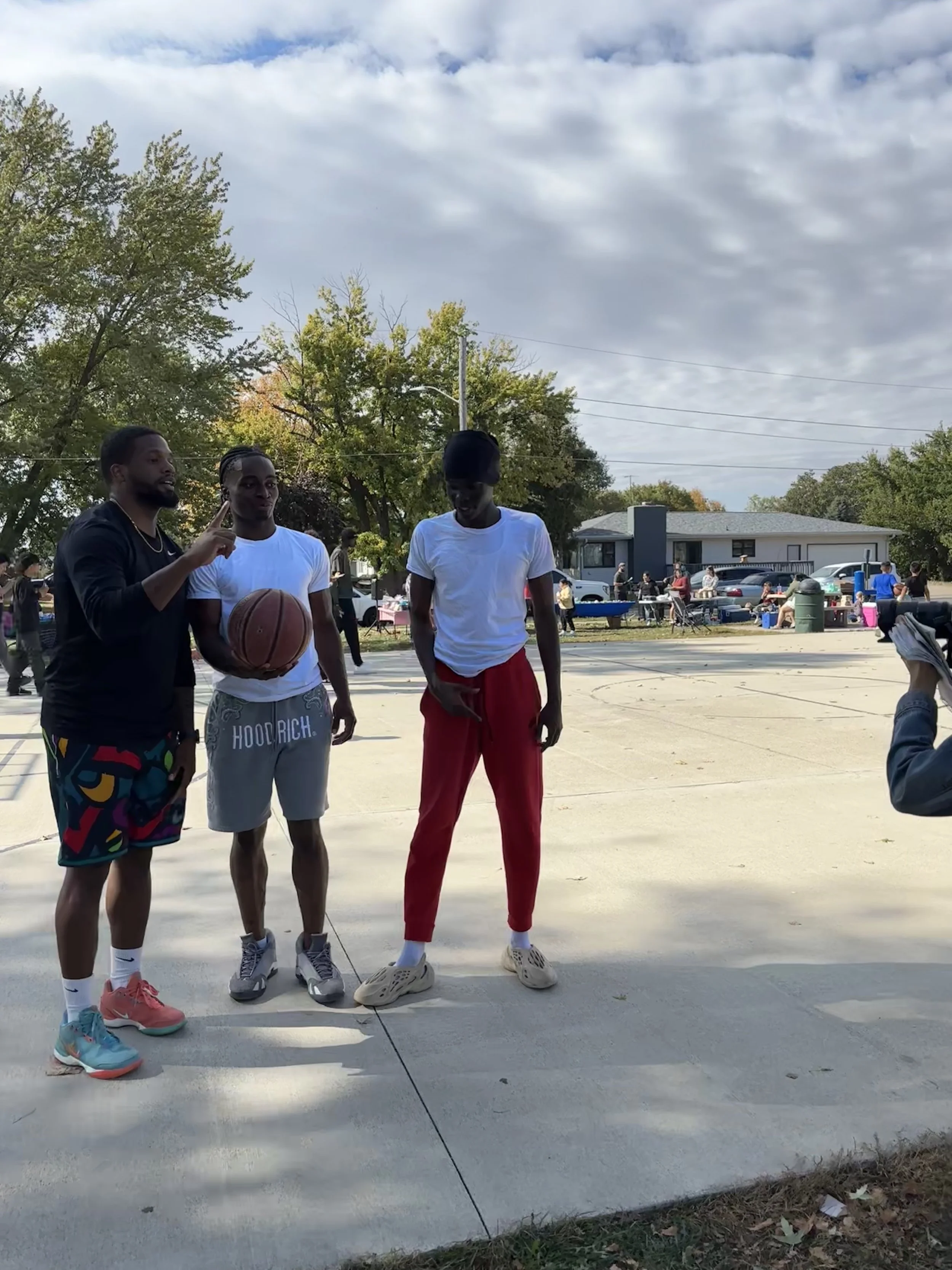 Three young men standing on a basketball court outdoors, one holding a basketball, with a group of people and tables in the background, under a partly cloudy sky.