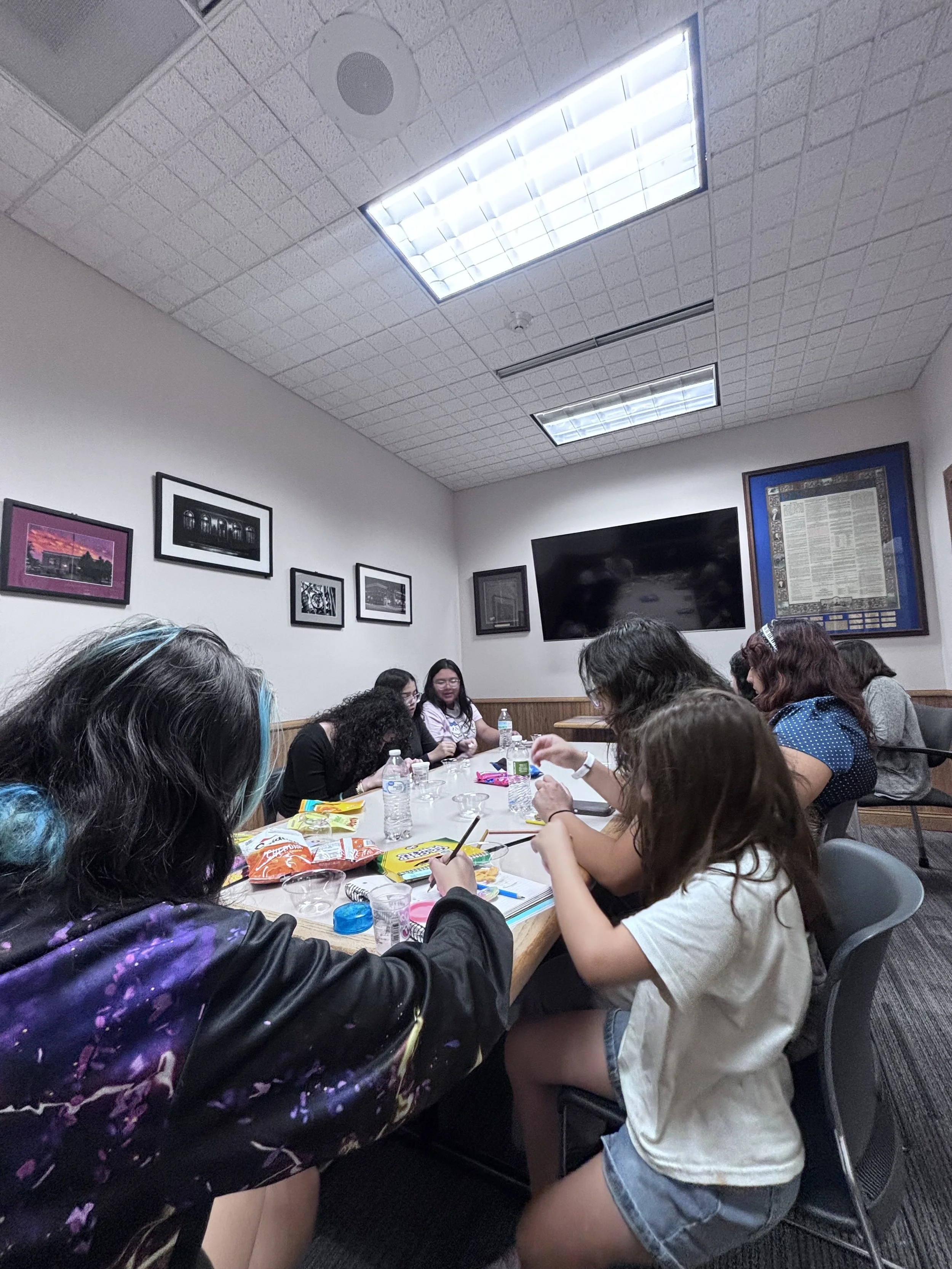 A group of people sitting around a large table in a conference room or classroom, engaging in discussion or activities. The table has snacks, water bottles, and papers. The room has framed pictures on the wall, a large television screen, and ceiling lights.