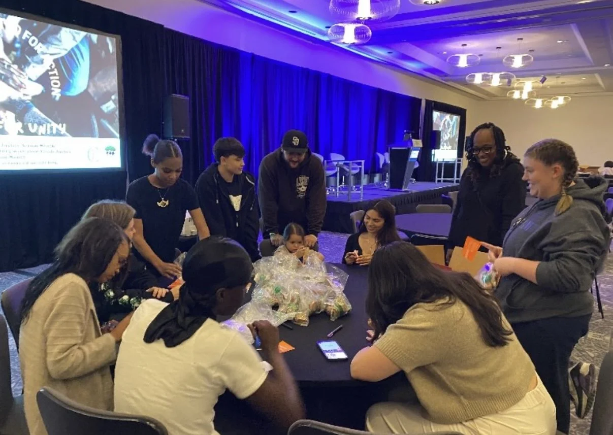 A group of people, including children and adults, gathered around a table in a conference room, engaging in a hands-on activity with various items on the table, with a stage and large screens in the background.