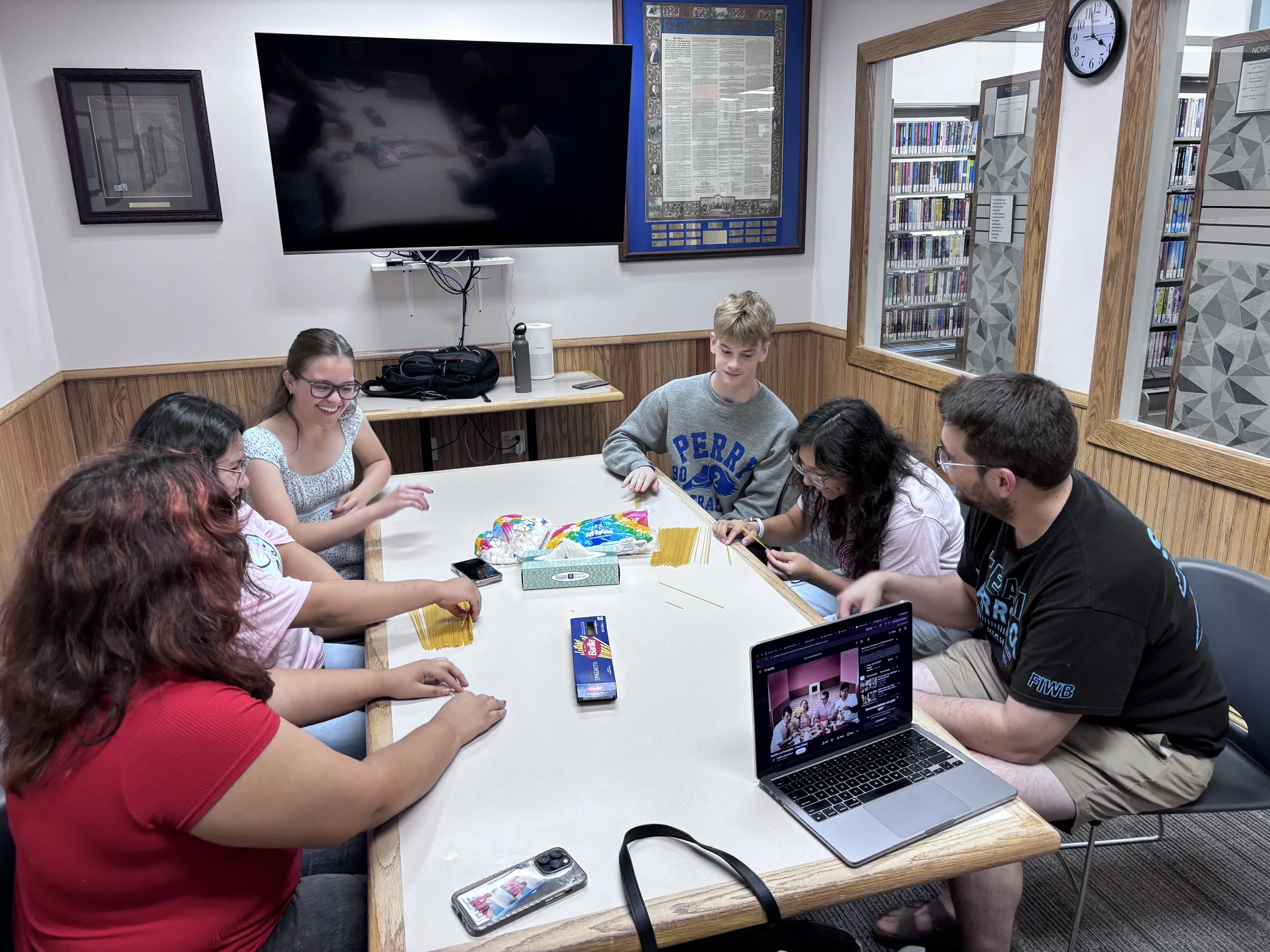 A group of young people sitting around a rectangular table in a library or study room, engaging in a multi-generational activity. They are discussing and working on something together, with materials like spaghetti and a box of tissues on the table. There is a large TV, a framed picture, and bookshelves behind them, along with a clock on the wall. Laptops and smartphones are also visible on the table.