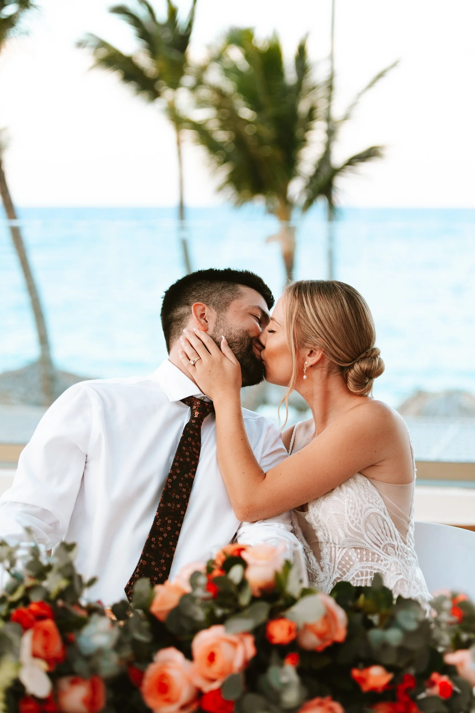A couple is kissing at a wedding reception with a view of the ocean and palm trees in the background, decorated with a floral arrangement of pink and red roses.