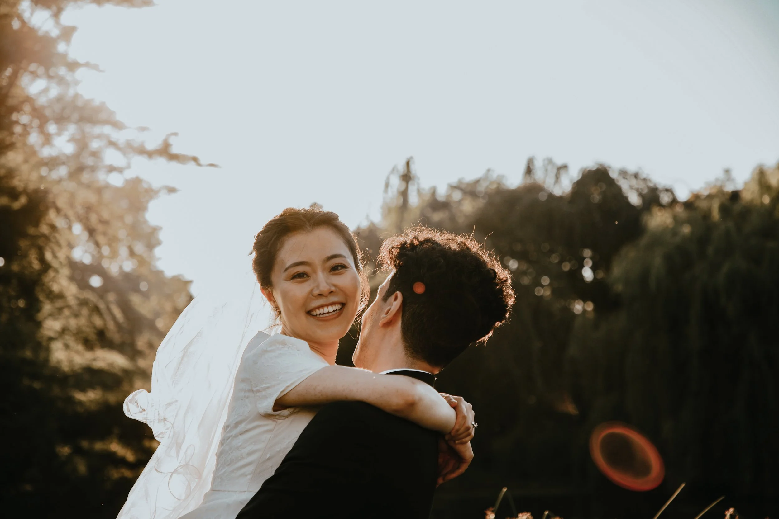 A bride with a white dress and veil is being lifted in the air by a groom in a black suit, outside during sunset with trees in the background.