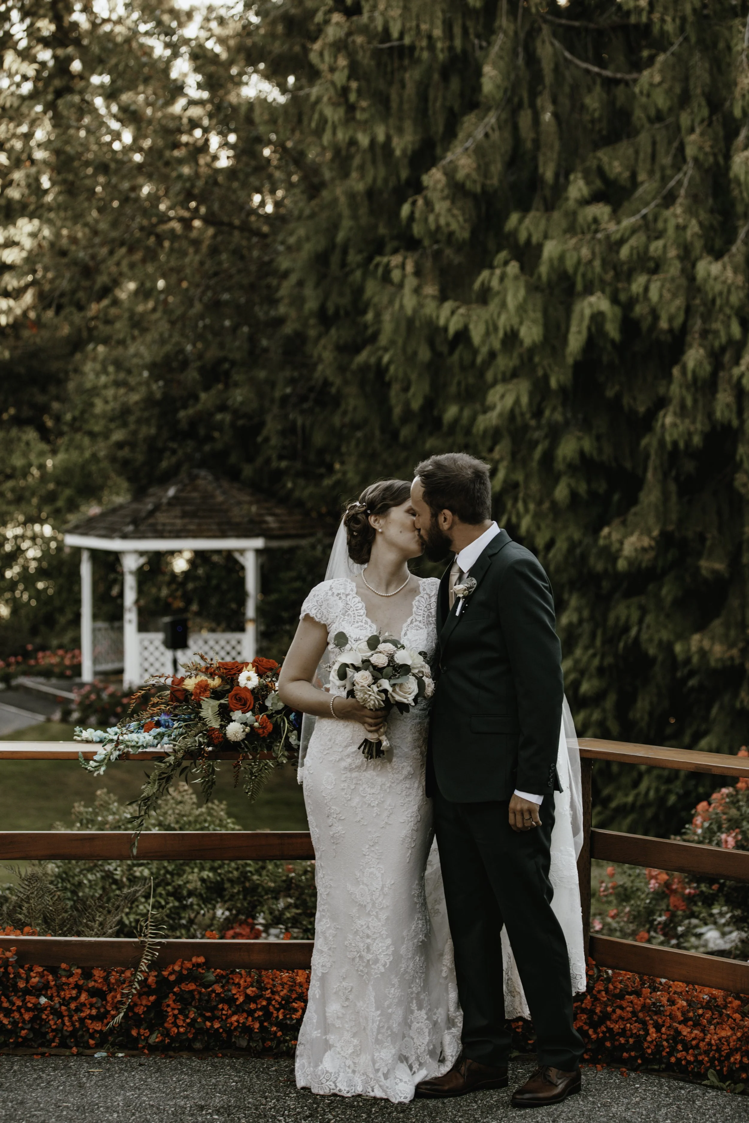 A bride and groom kiss on a wooden bridge during their wedding, with a bouquet in the bride's hand and colorful flowers behind them.