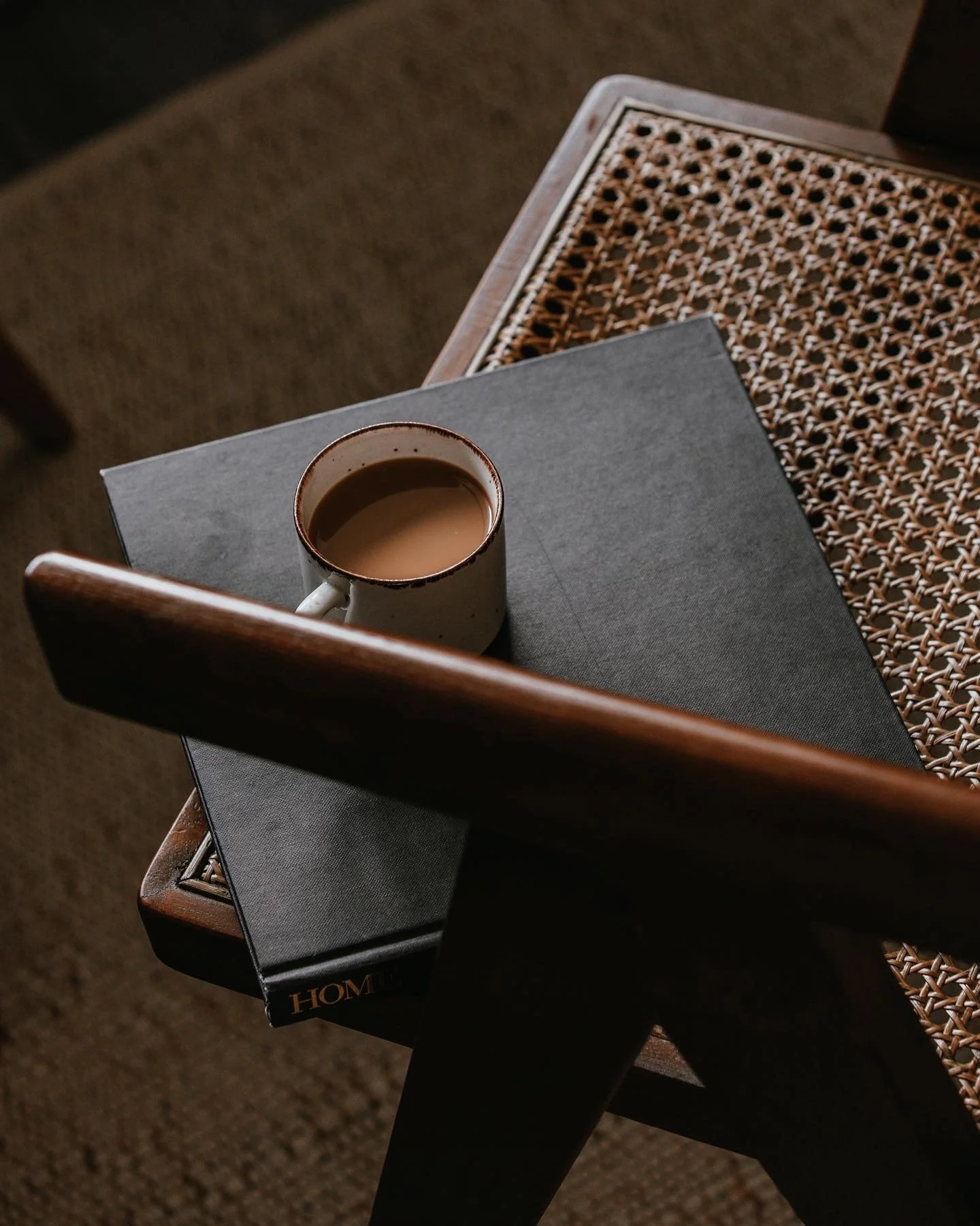 A cup of coffee with cream on a gray book, placed on a black tabletop, with a rattan chair and brown carpet in the background.