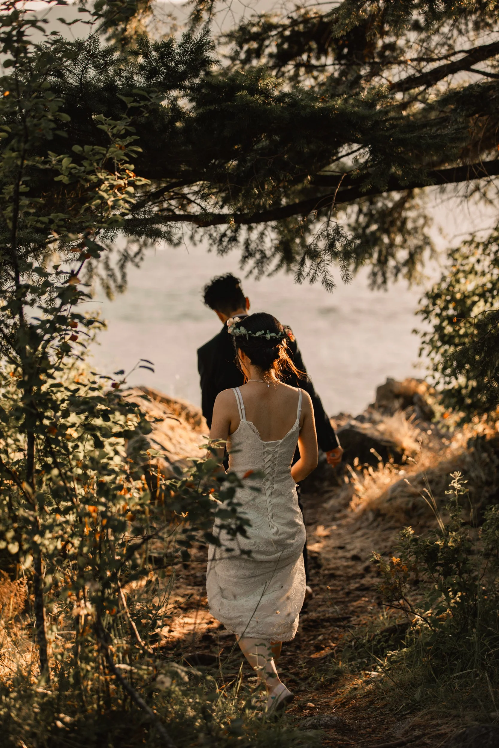 A bride and groom walking along a rocky path near the water, surrounded by trees, during sunset.