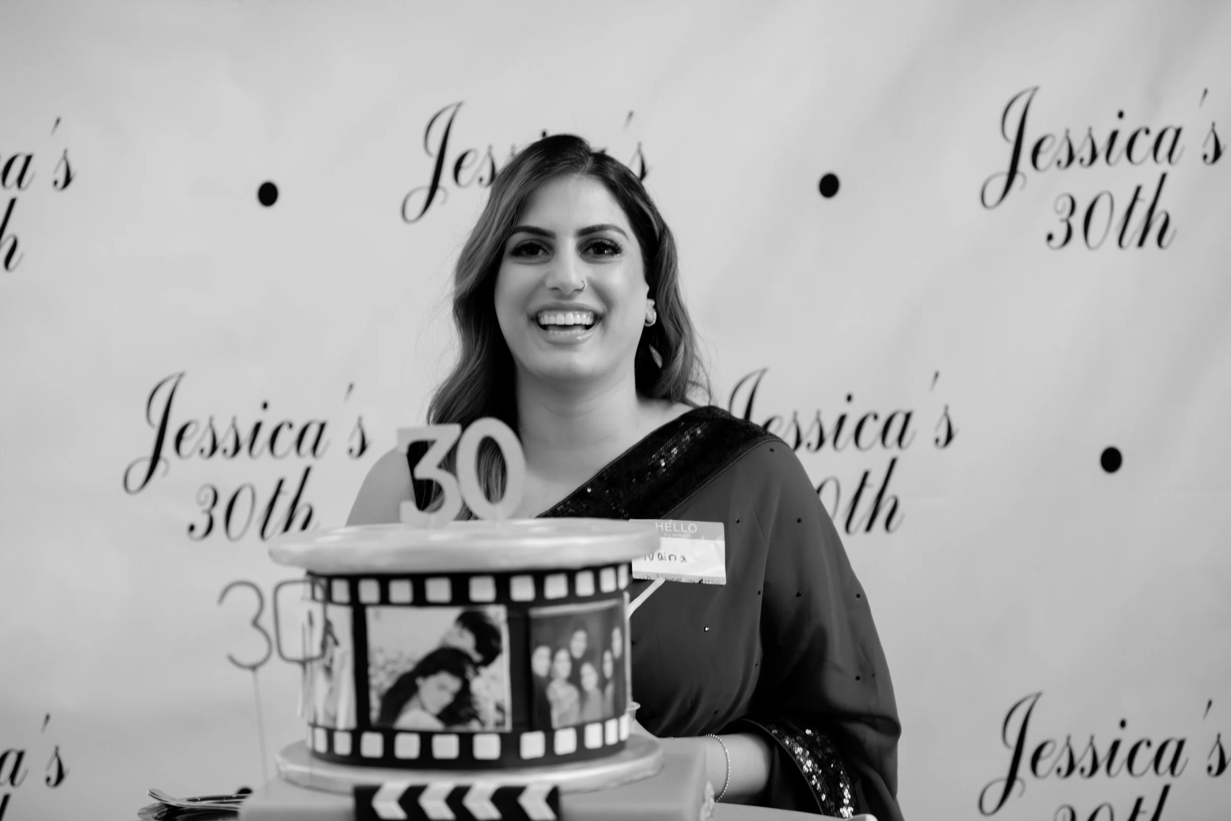 A woman celebrating her 30th birthday at an event with a cake decorated with photos and the number 30. The backdrop reads "Jessica's 30th." She is smiling and wearing a dark outfit with sequin details.