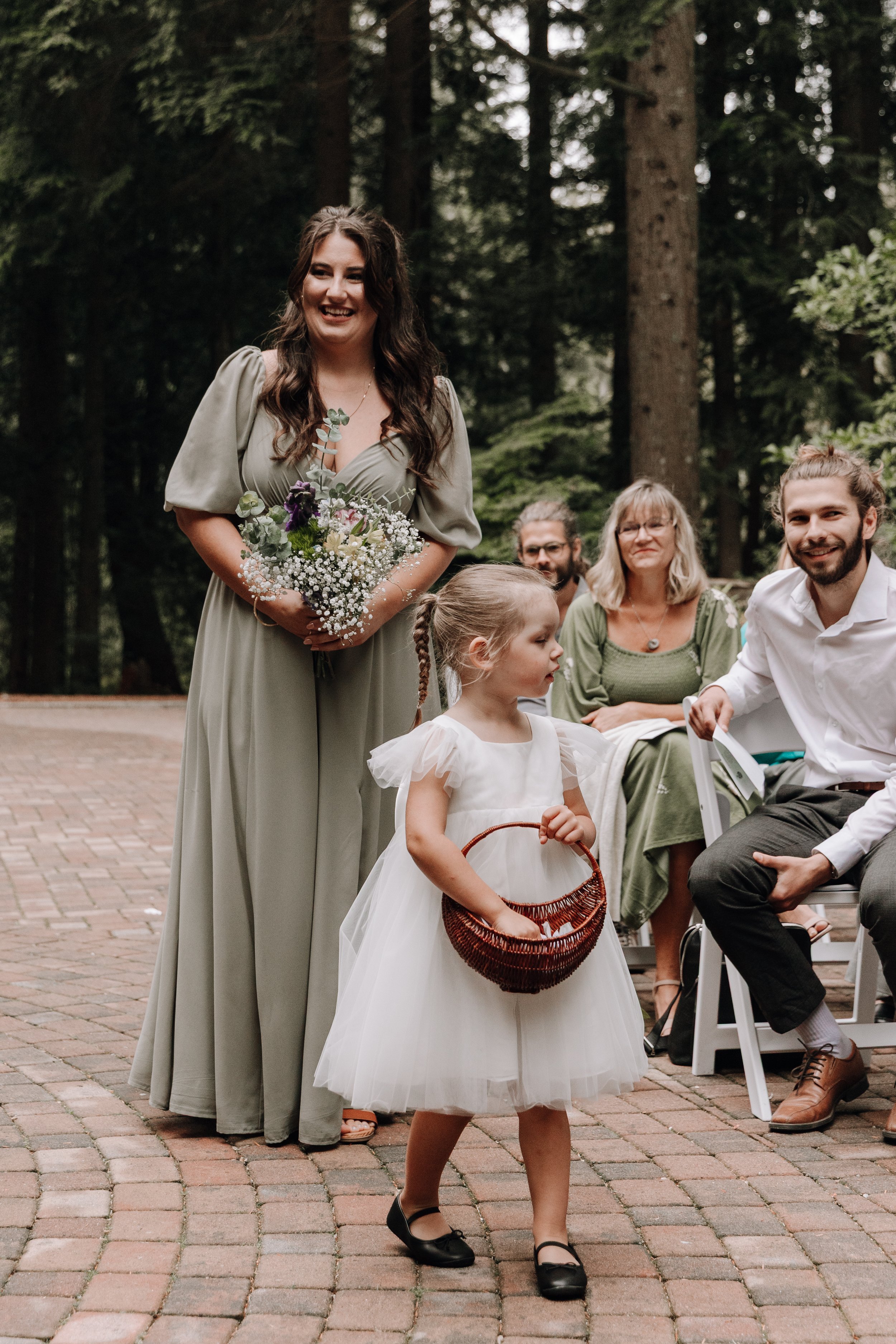 A young girl in a white dress holding a wicker basket, walking outdoors on a brick pathway, with a woman in a gray dress holding a bouquet of flowers and smiling, and family members seated in chairs nearby, with trees in the background.
