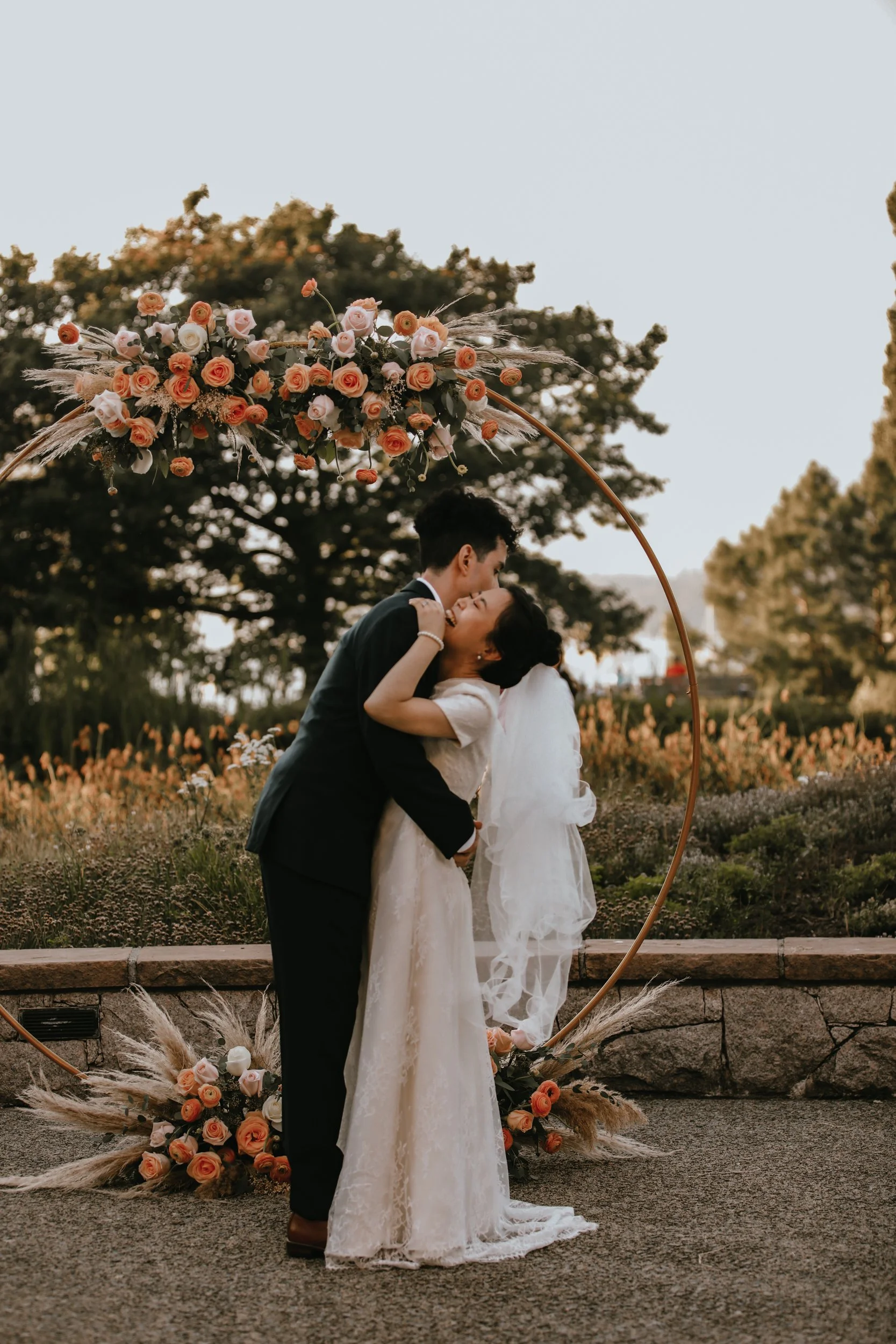 A bride and groom are embracing in front of a floral wedding arch outdoors during sunset.