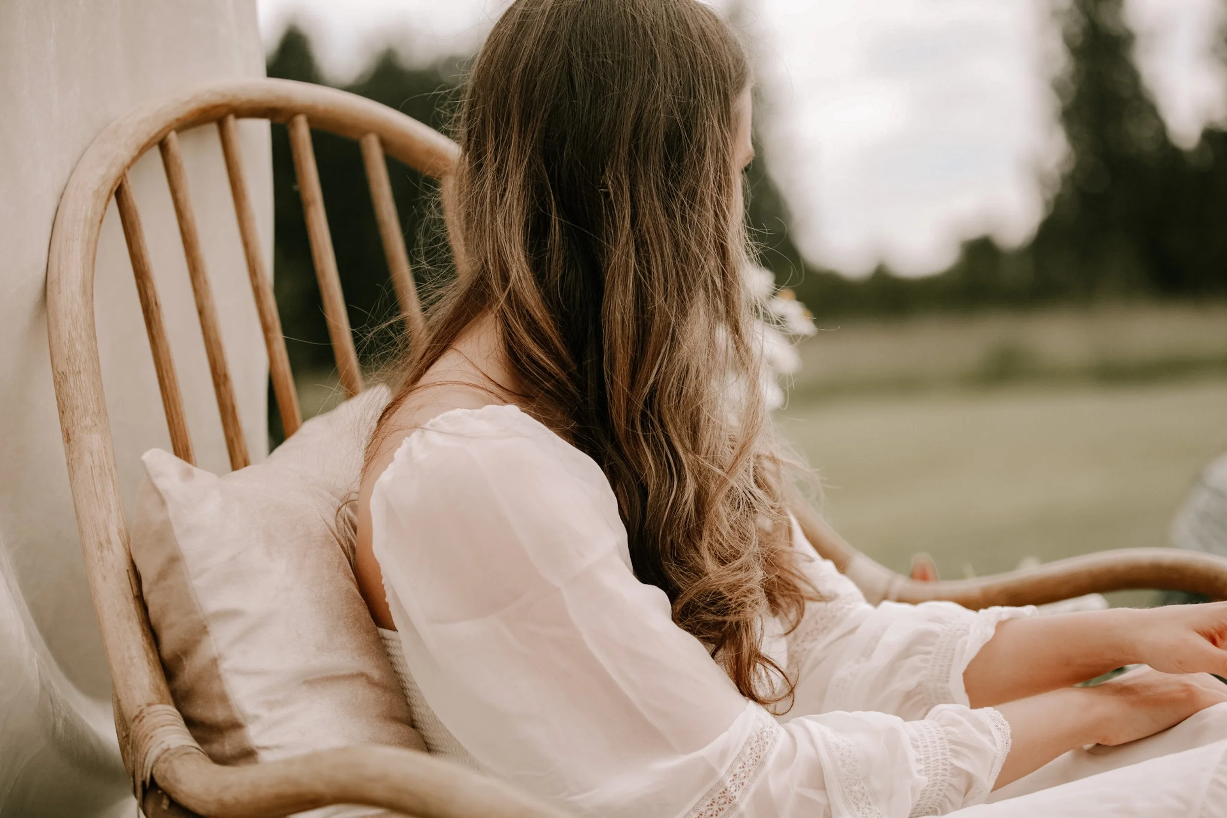 A woman with long, wavy hair sitting on a wooden bed outdoors, facing away, with a pillow behind her back, wearing a white dress, with a blurred landscape in the background.
